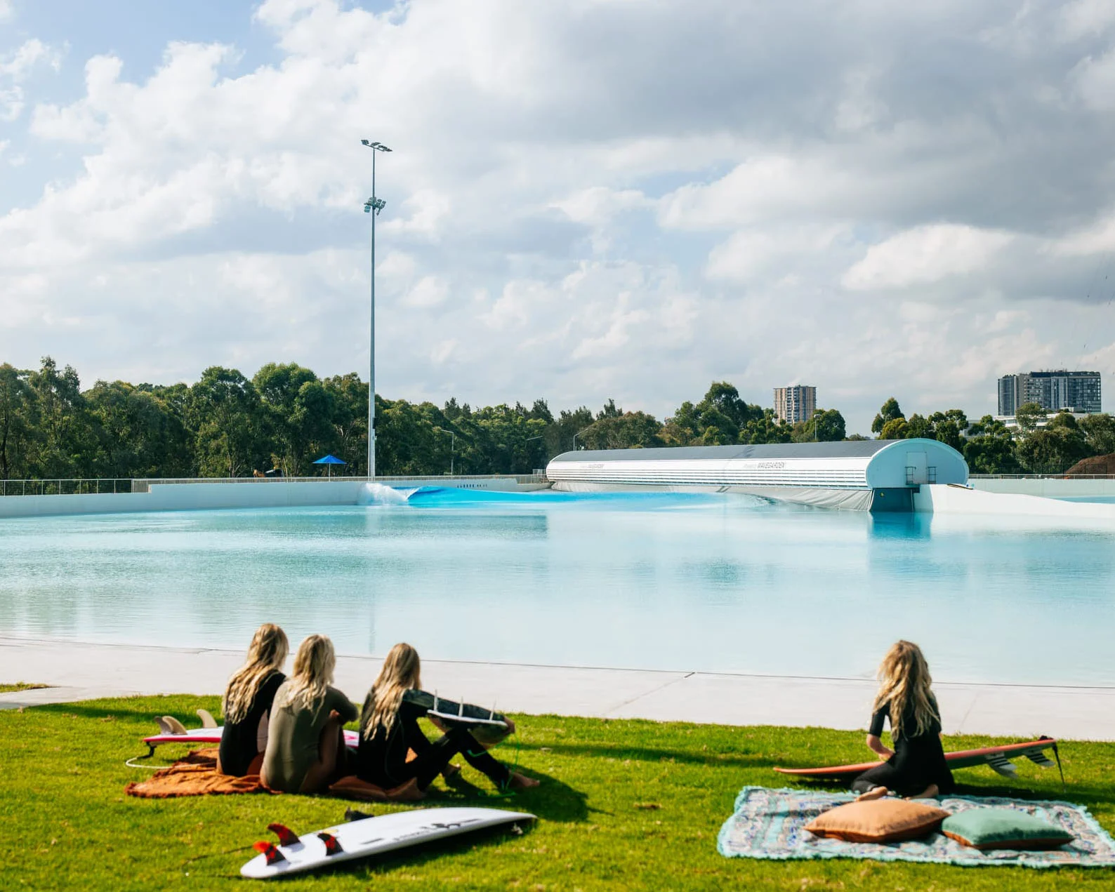 A photograph of four people sitting on grass next to surfboards and a large wave pool.