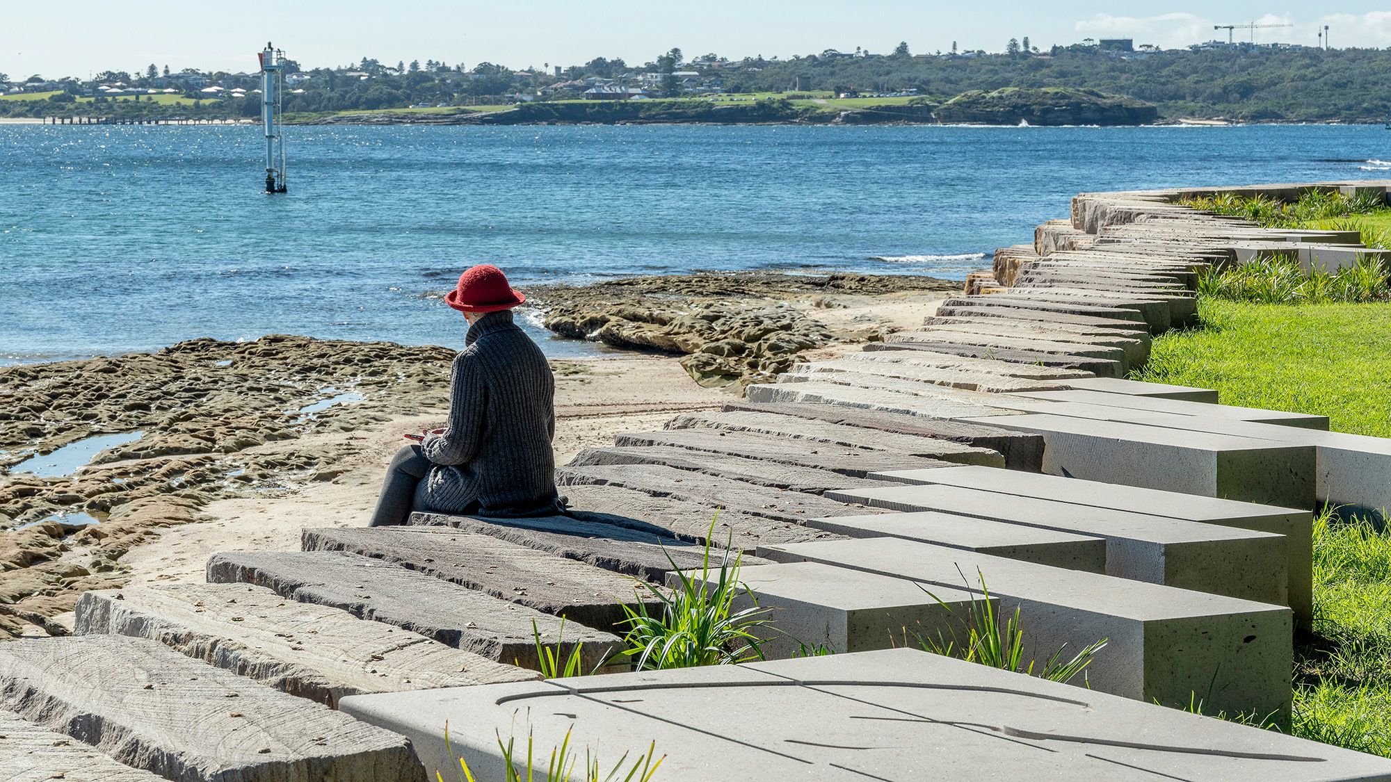 Woman sits on the block forming part of revetment wall and looks out on the bay.
