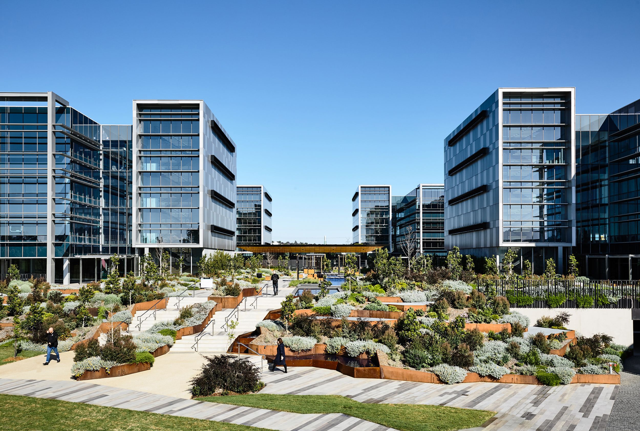 A photograph of a new business park with glass office buildings to the left and right. The space between is filled with a mix of plants in core ten steel planters. A staircase leads into the site. 