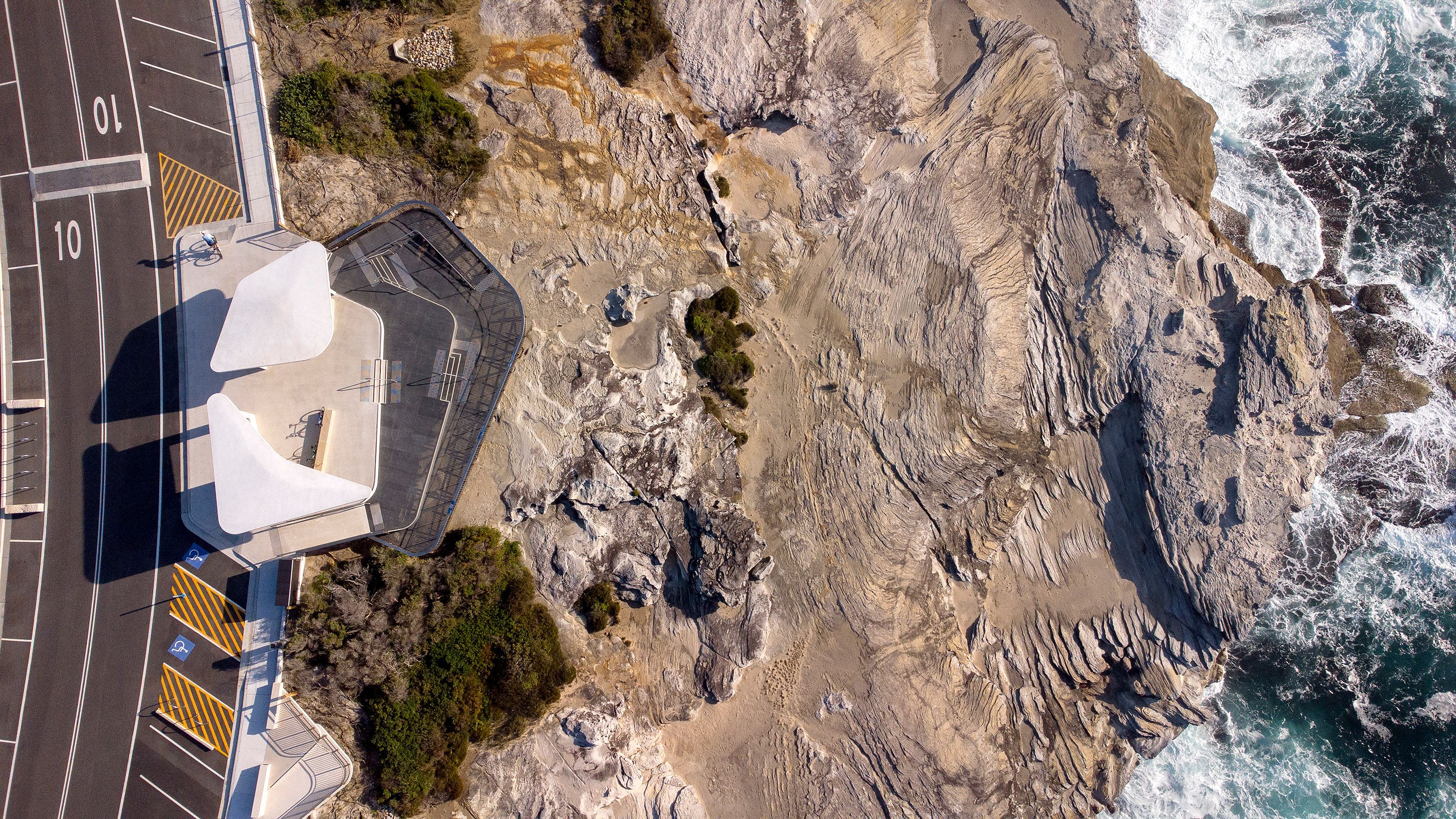 An aerial photograph of a sandstone coastline. On the left a contemporary white viewing platform and road is visible. On the right of the image, waves crash against cliffs. 