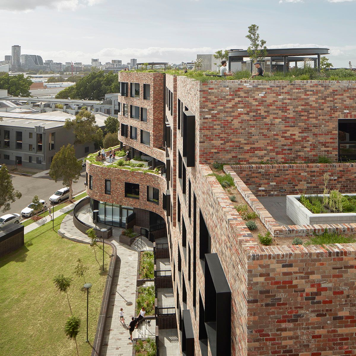A photograph of a brick apartment building next to a pocket park, with a rooftop garden, and vegetable gardens on terraces.  