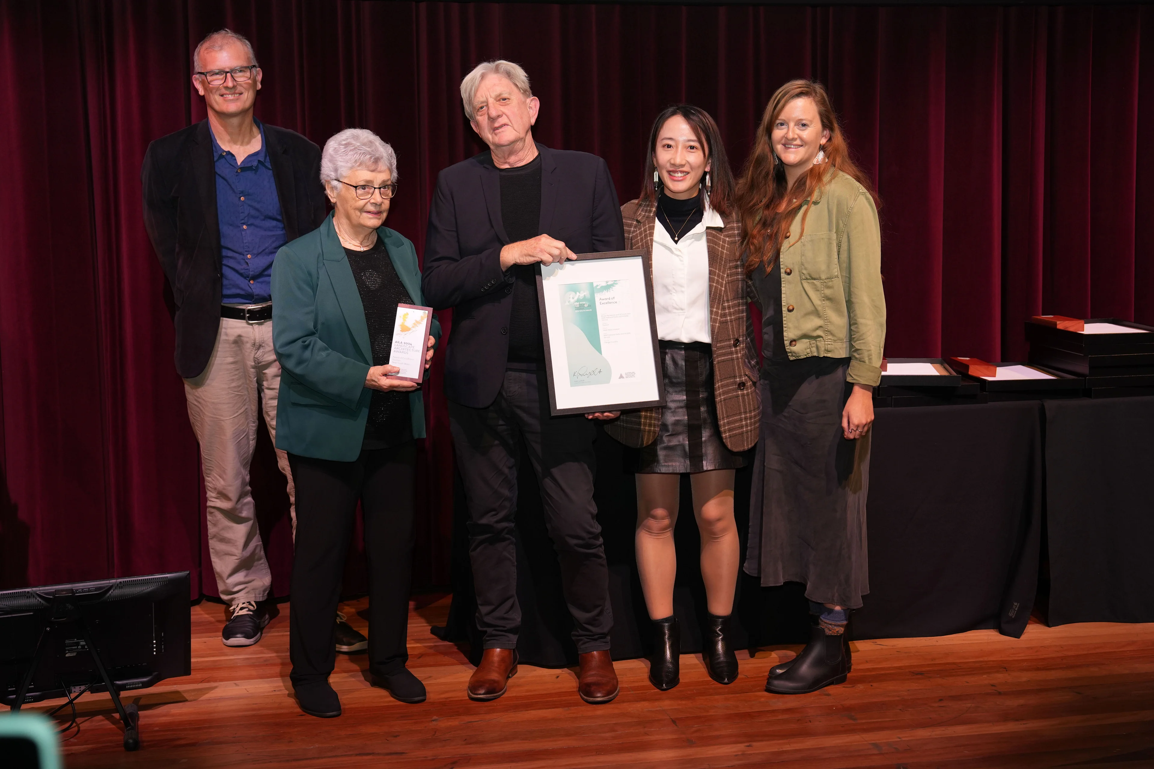 A photo of five people on stage accepting an award. One person holds a trophy, one person holds a framed certificate.