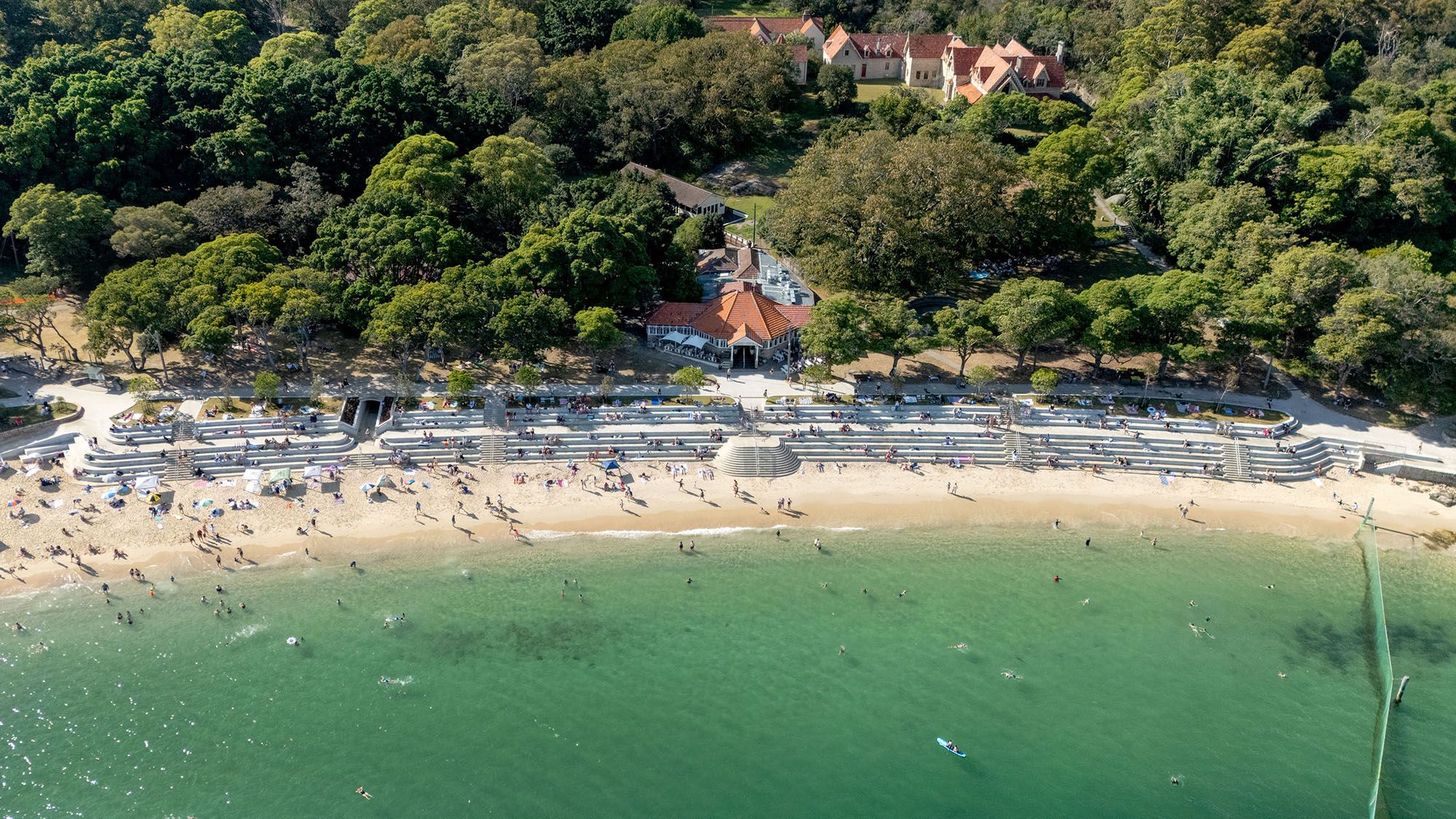 A drone photograph looking down on a sunny beach with a tiered seawall. There are heritage buildings near the water.