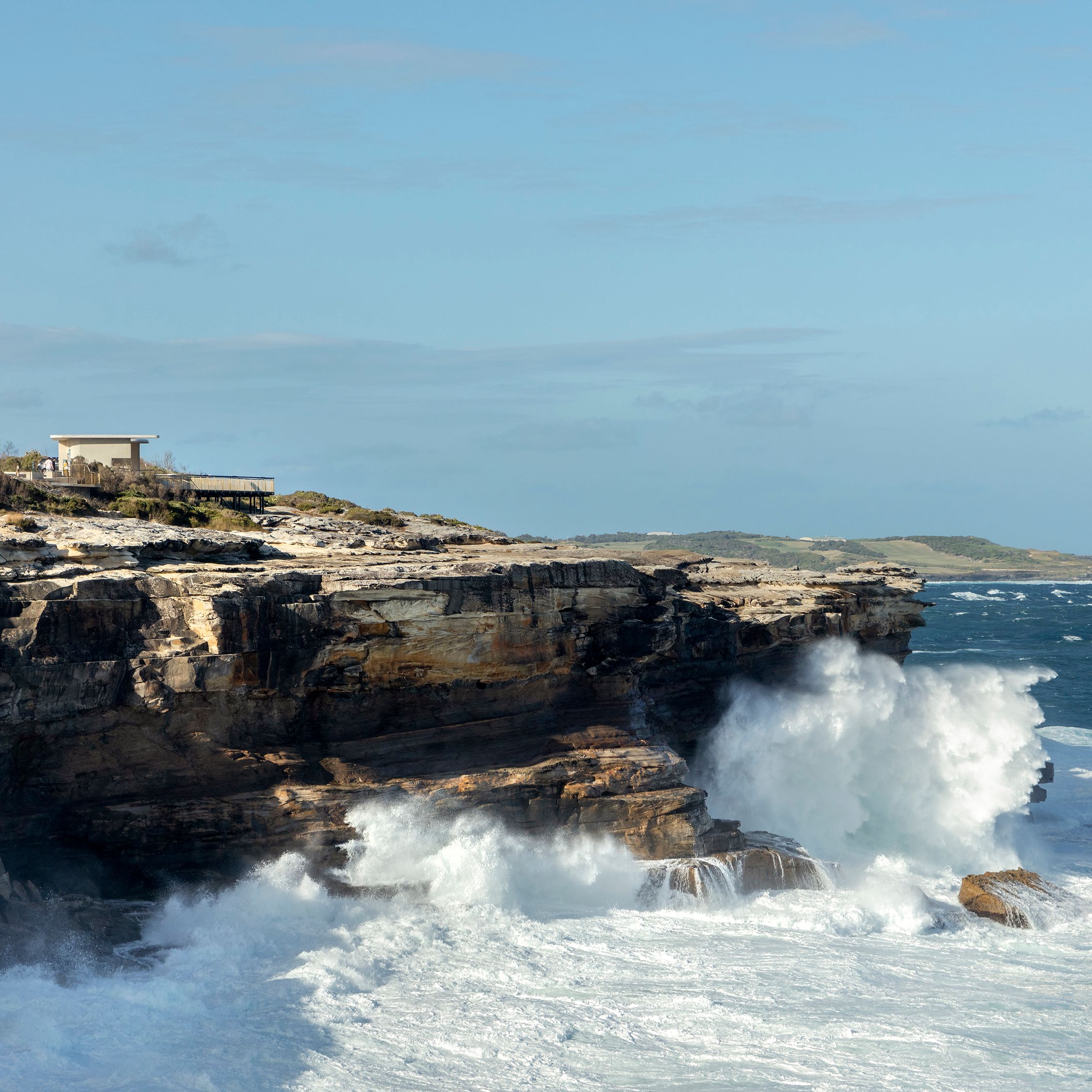 A photograph of large ocean waves crashing against a sandstone cliff, the sky is blue with scattered clouds, and there is a viewing platform blending into the landscape on the top of the cliff. 