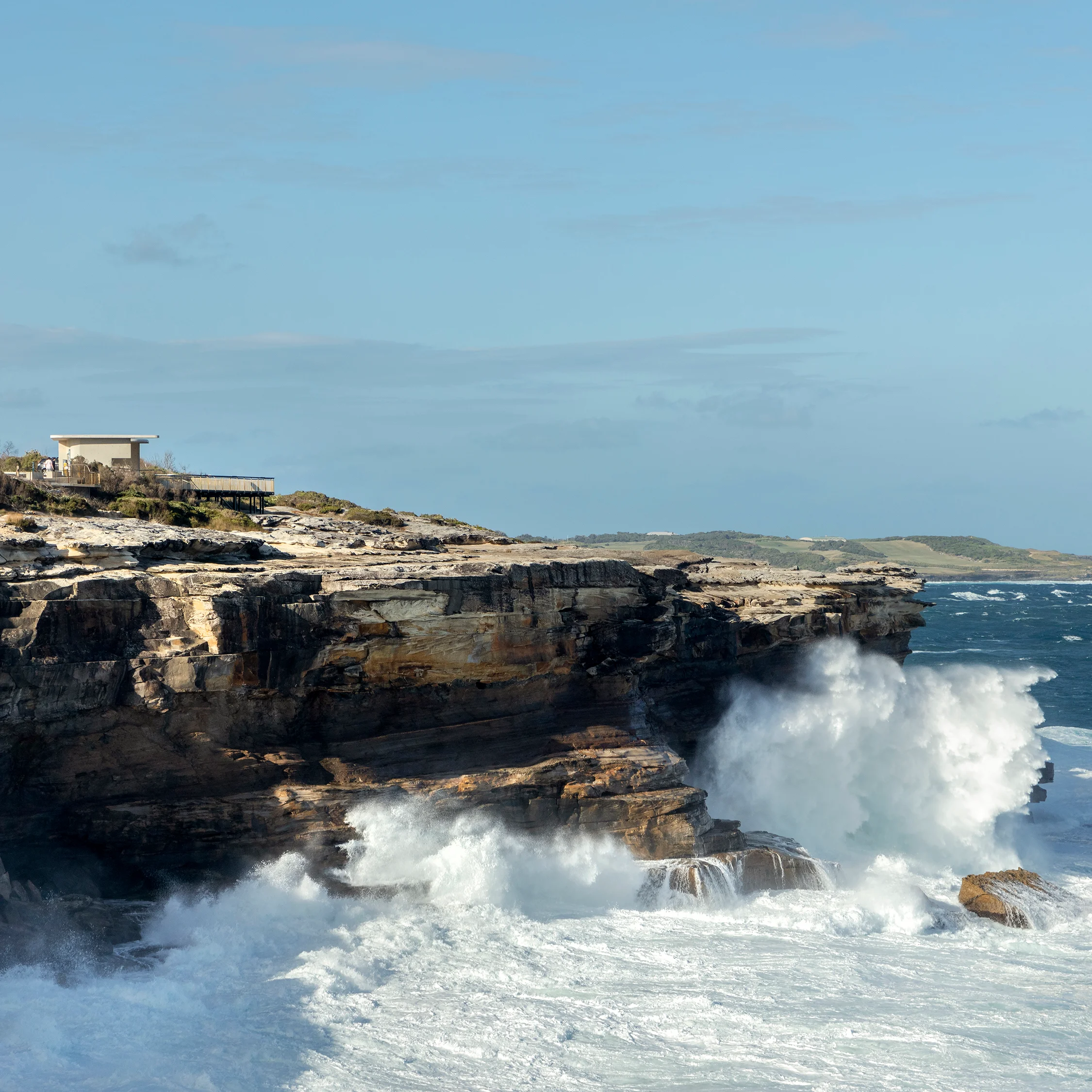 A photograph of large ocean waves crashing against a sandstone cliff, the sky is blue with scattered clouds, and there is a viewing platform blending into the landscape on the top of the cliff.