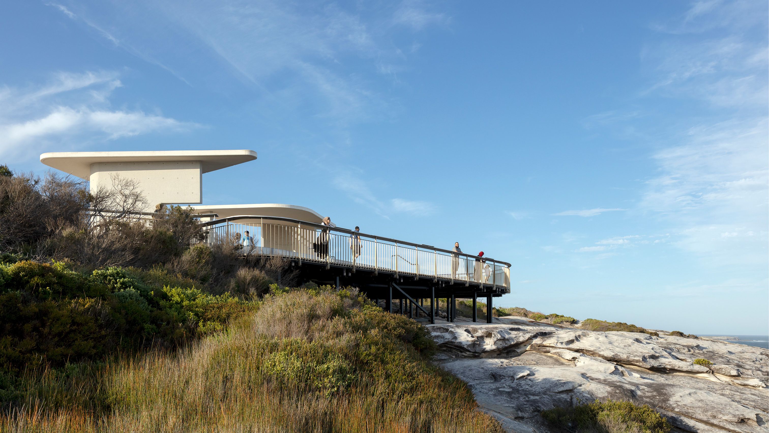 A photograph of a viewing platform taken from a low angle with a blue sky above. A few people are leaning against the railing and looking out. Vegetation and rocks are seen in the foreground. 