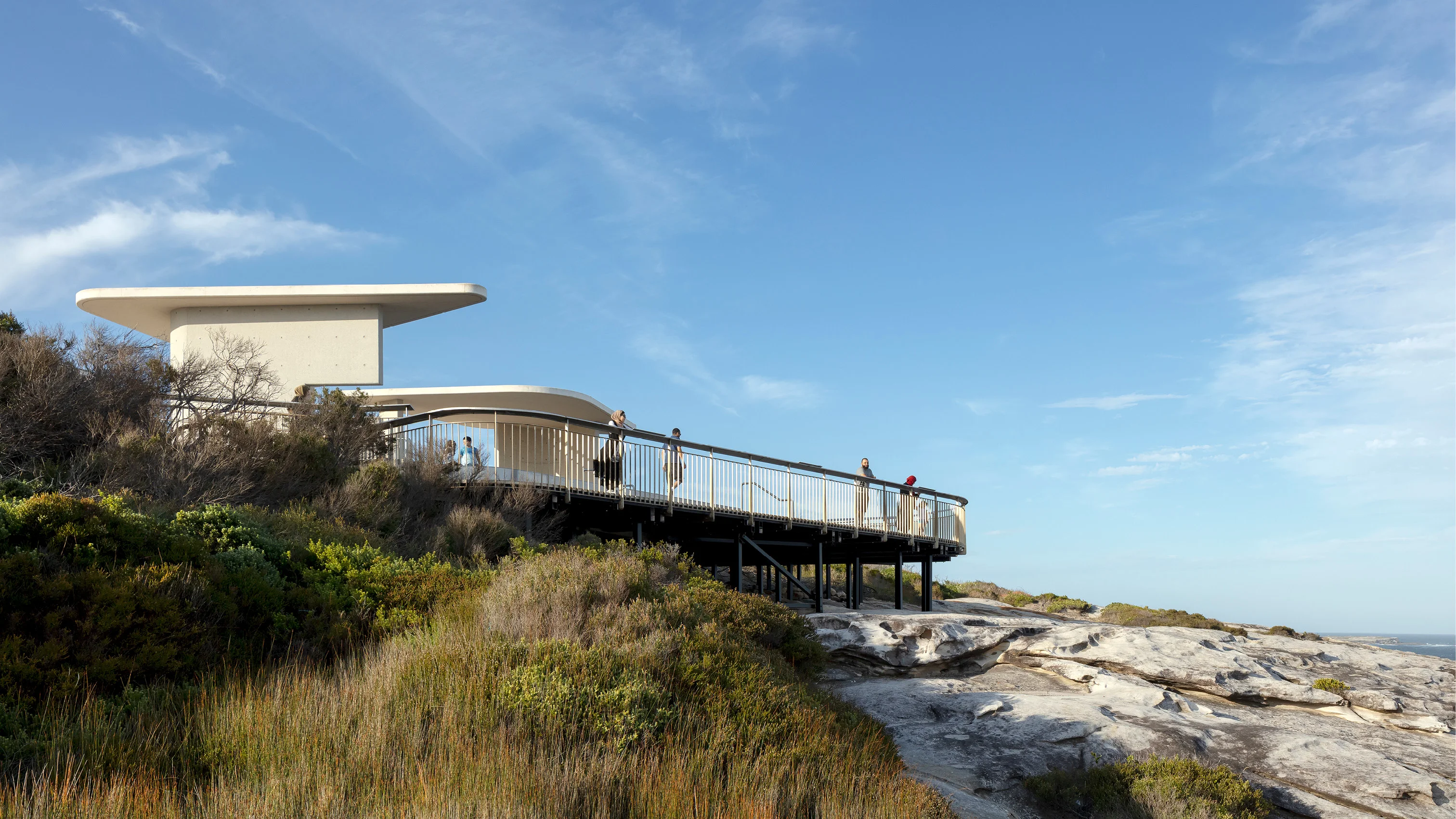 A photograph of a viewing platform taken from a low angle with a blue sky above. A few people are leaning against the railing and looking out. Vegetation and rocks are seen in the foreground.