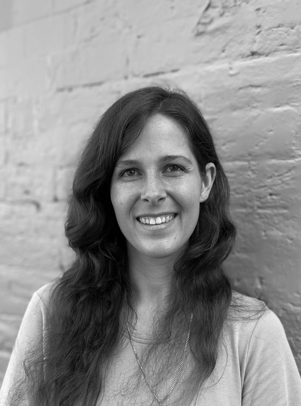 A black and white headshot of a smiling person in front of a brick wall.