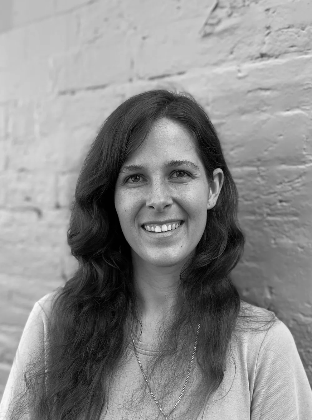 A black and white headshot of a smiling person in front of a brick wall.