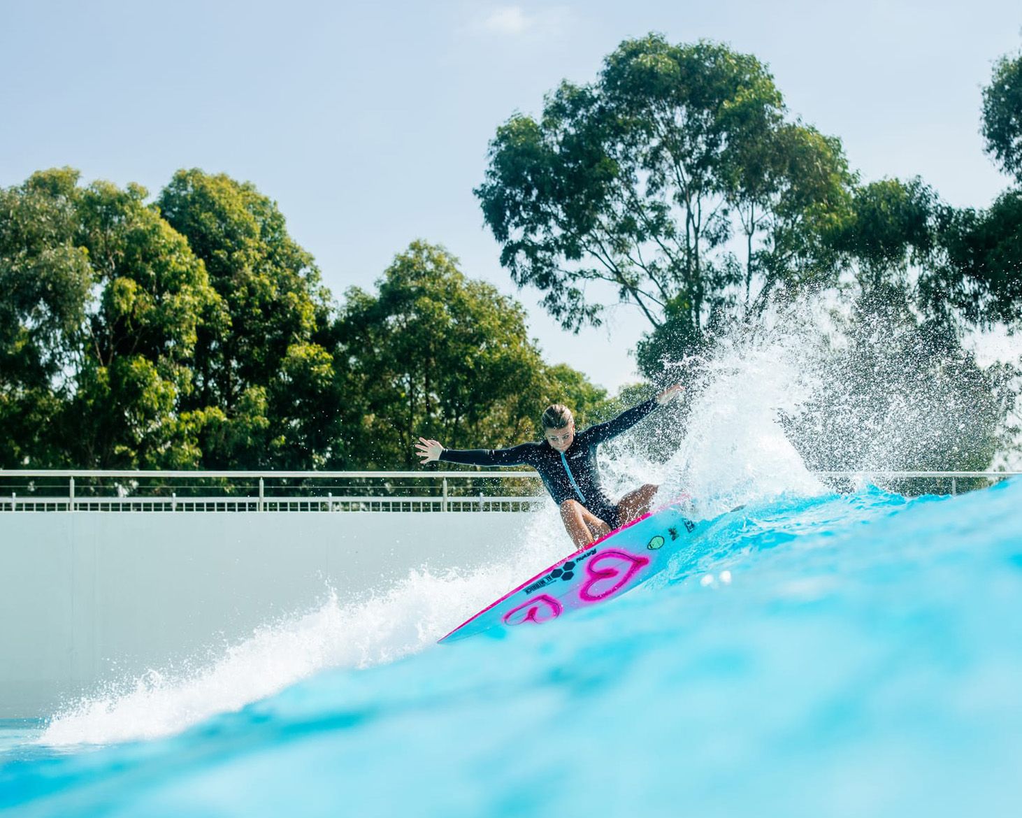 A photo of a surfer riding in a wave pool. There are two pink hearts spray painted on their surfboard.