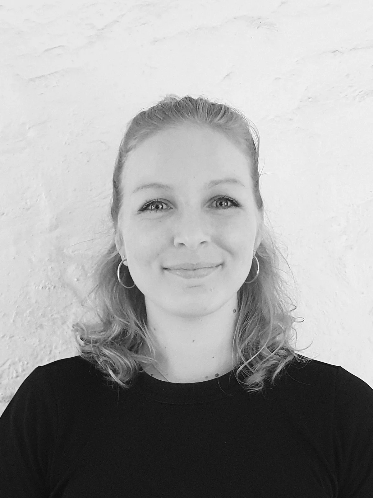 A black and white headshot of a person wearing black, standing in front of a whitewall.