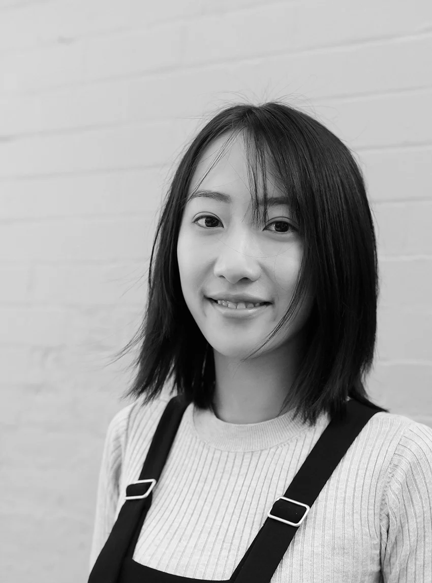 A black and white headshot of a person wearing black overalls, standing in front of a brick wall painted white.