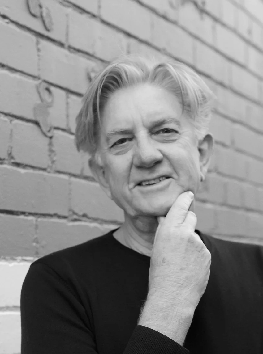 A black and white headshot of a smiling person in front of a brick wall.