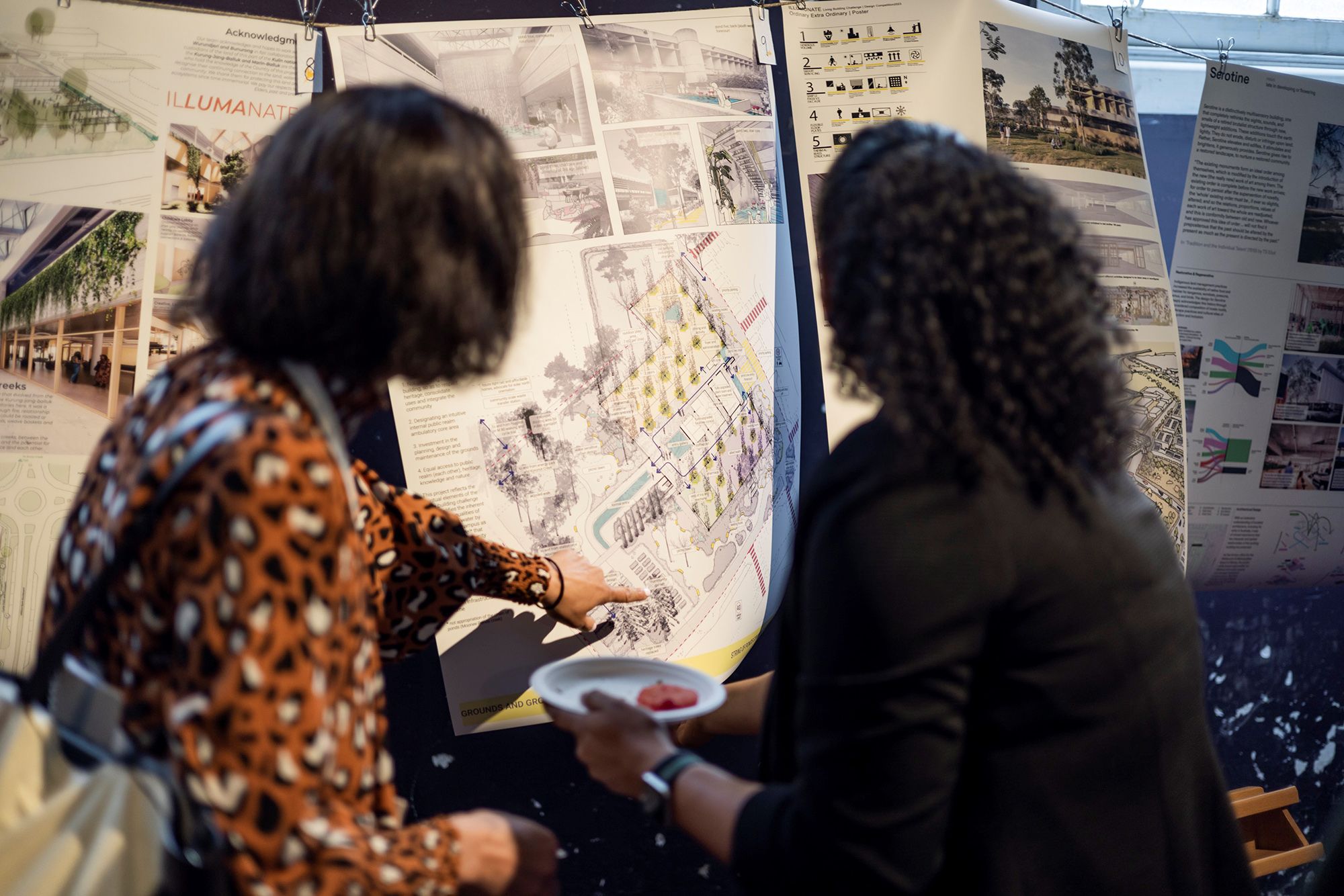 A photograph of two people from behind who are pointing at details on large posters with urban and architectural graphics.