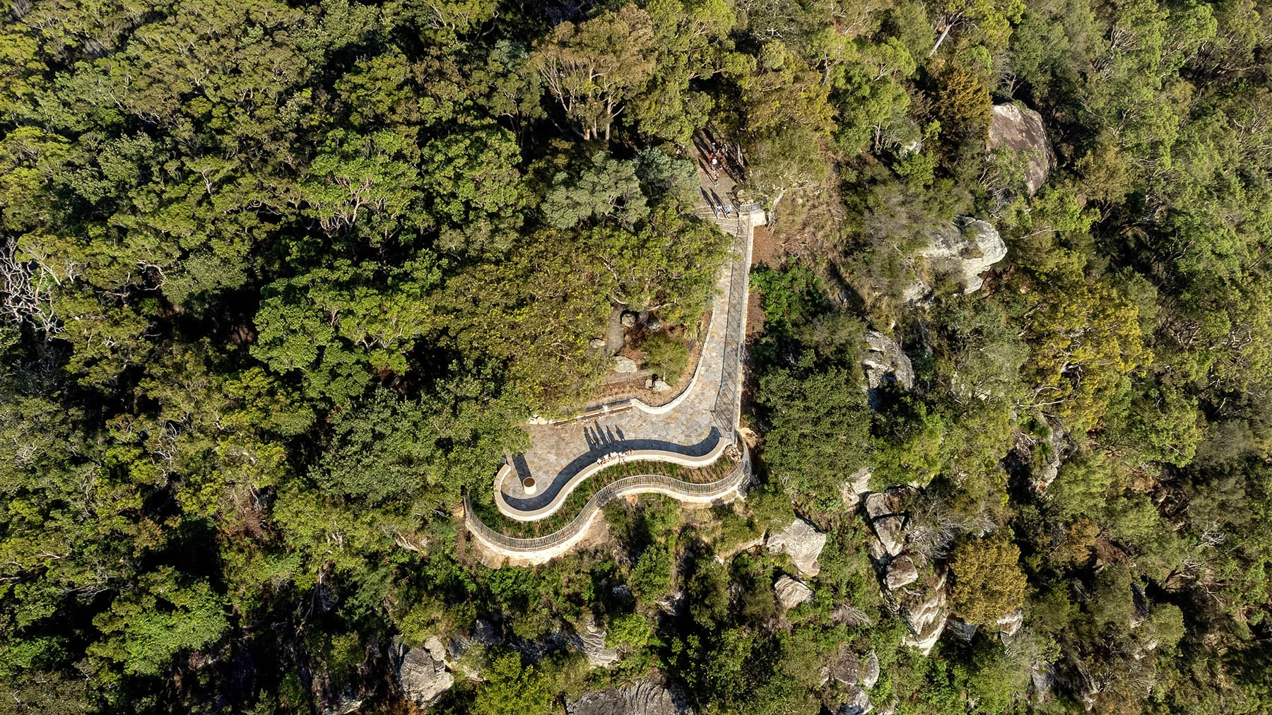 An aerial photograph of a curvy, sandstone lookout surrounded by green bushland, with people arriving and sitting on a low wall.