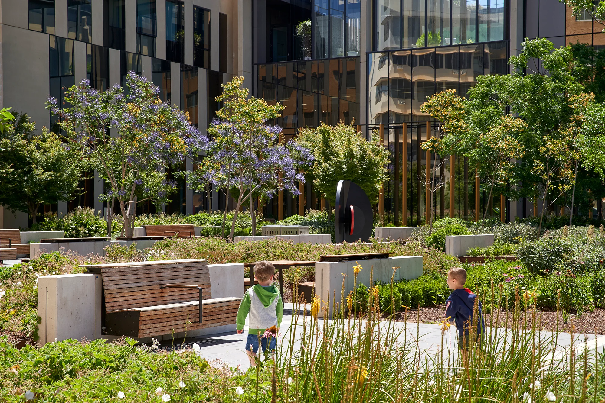 A photograph of two children playing in large public courtyard full of plants. There are various seating areas, with trees and large building facades in the background.