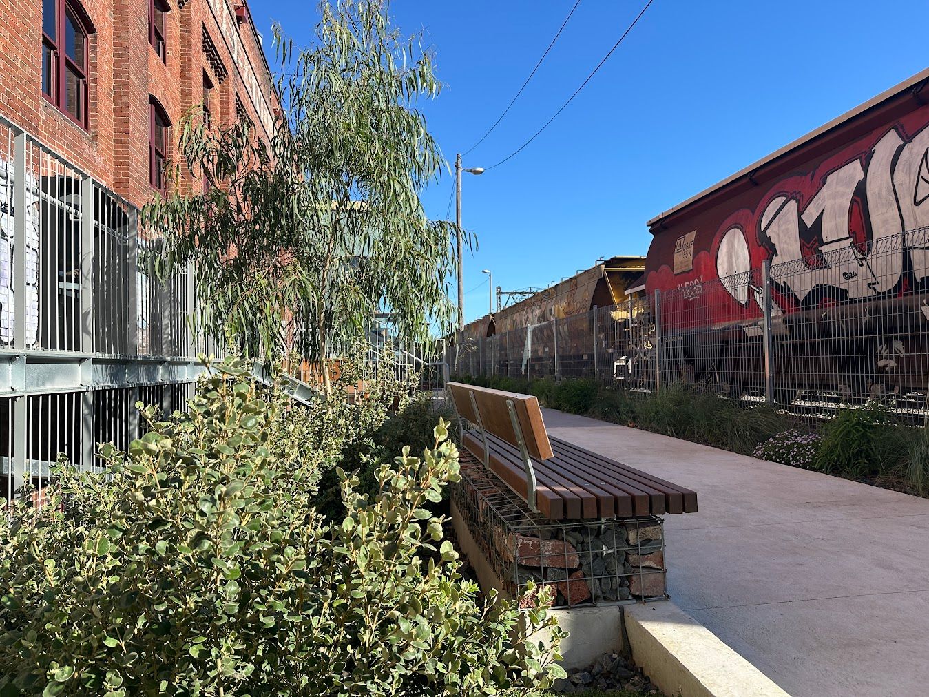 A photograph of pathways, planting, and seating between an old industrial building and railway line.