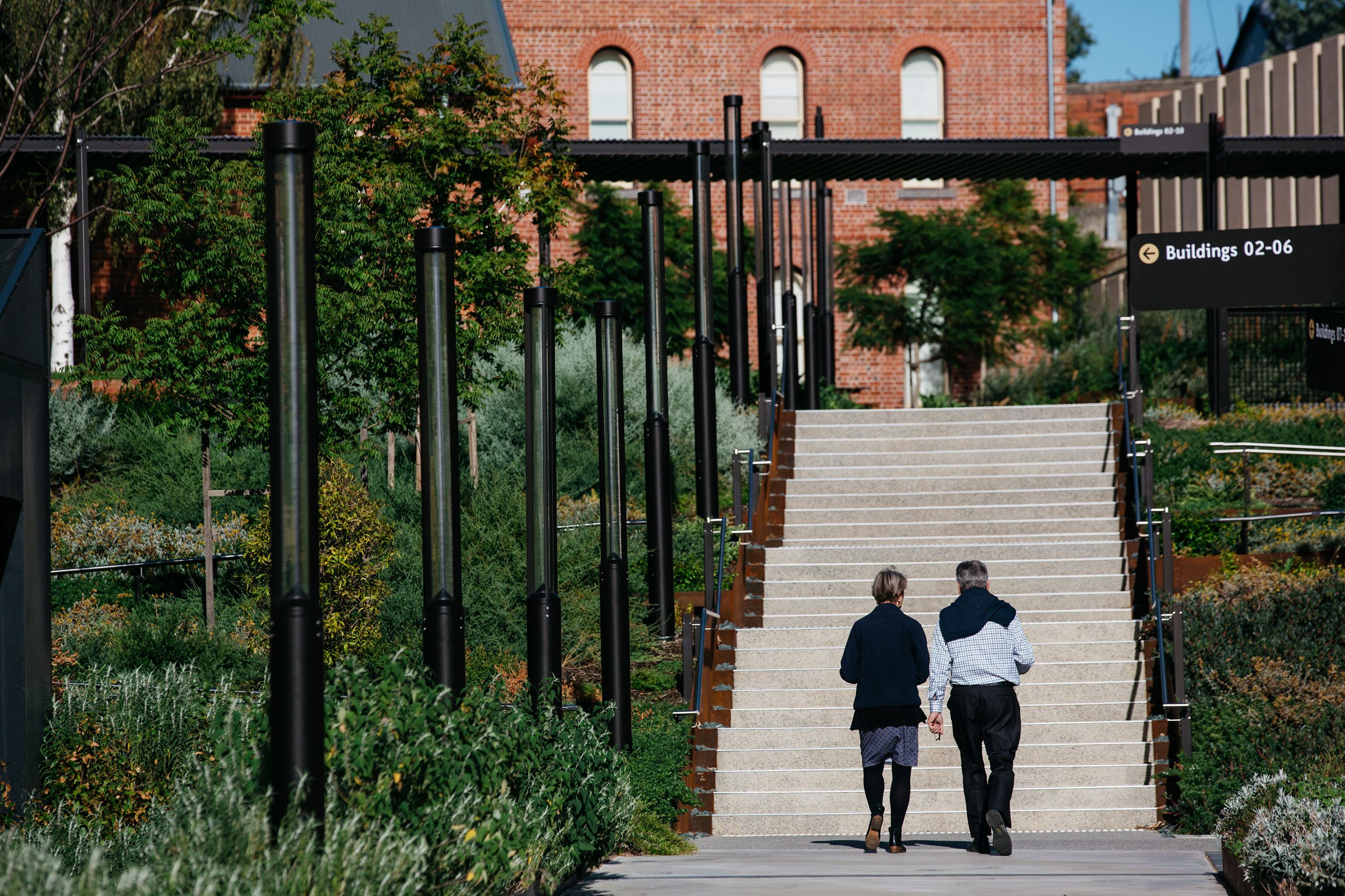 A photograph of two people walking towards an outdoor staircase, with plants either side. There is a red brick building at the top of the stairs, and black light fixtures to the left of their path.