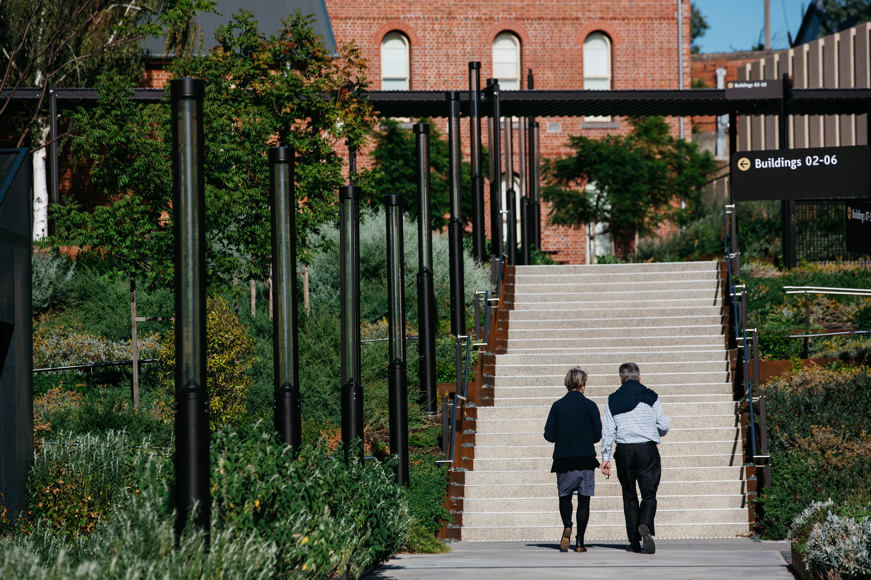 A photograph of two people walking towards an outdoor staircase, with plants either side. There is a red brick building at the top of the stairs, and black light fixtures to the left of their path.