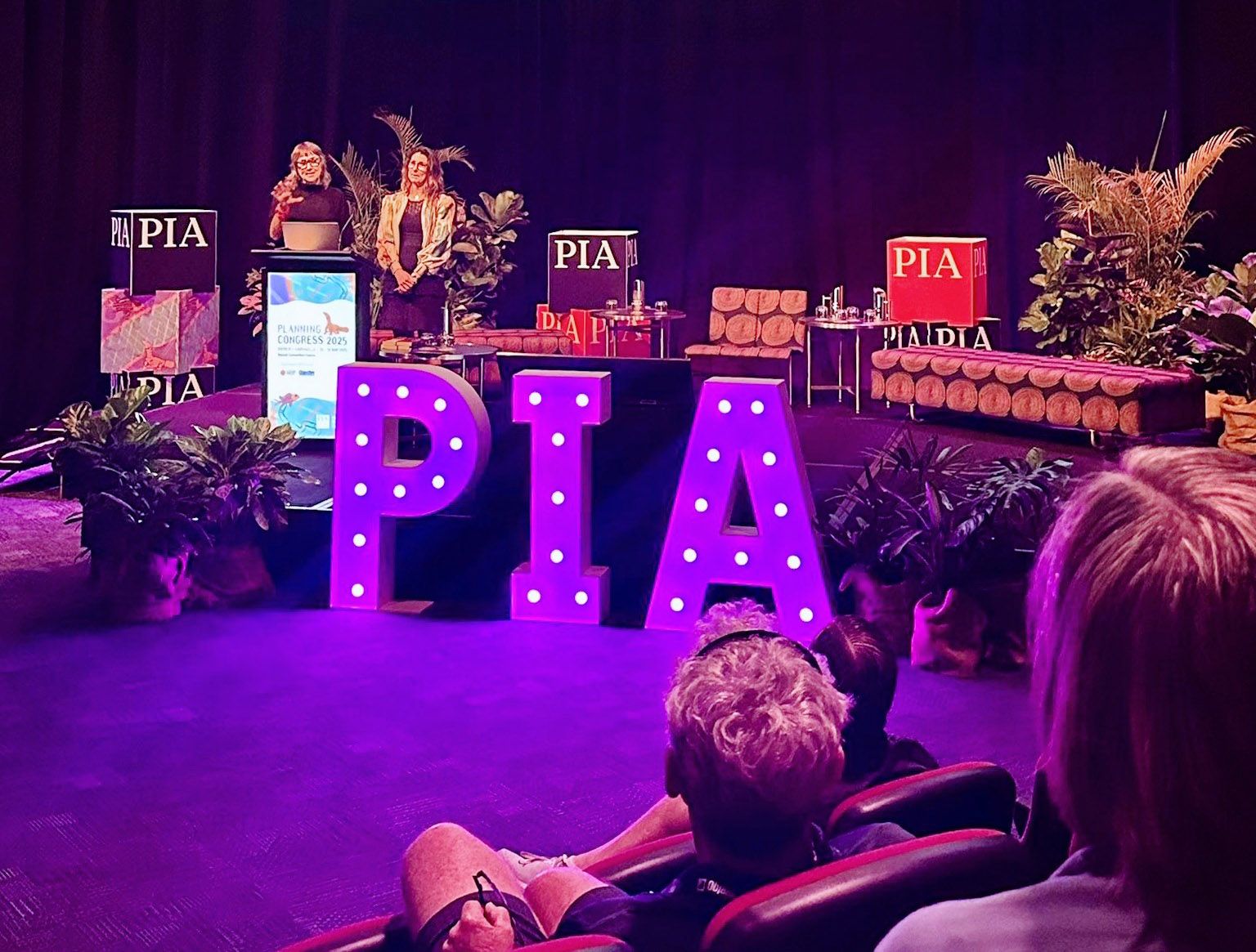A photo of two people presenting on a stage. There are flowers and "PIA" signs on the stage, and a purple glow across the audience.