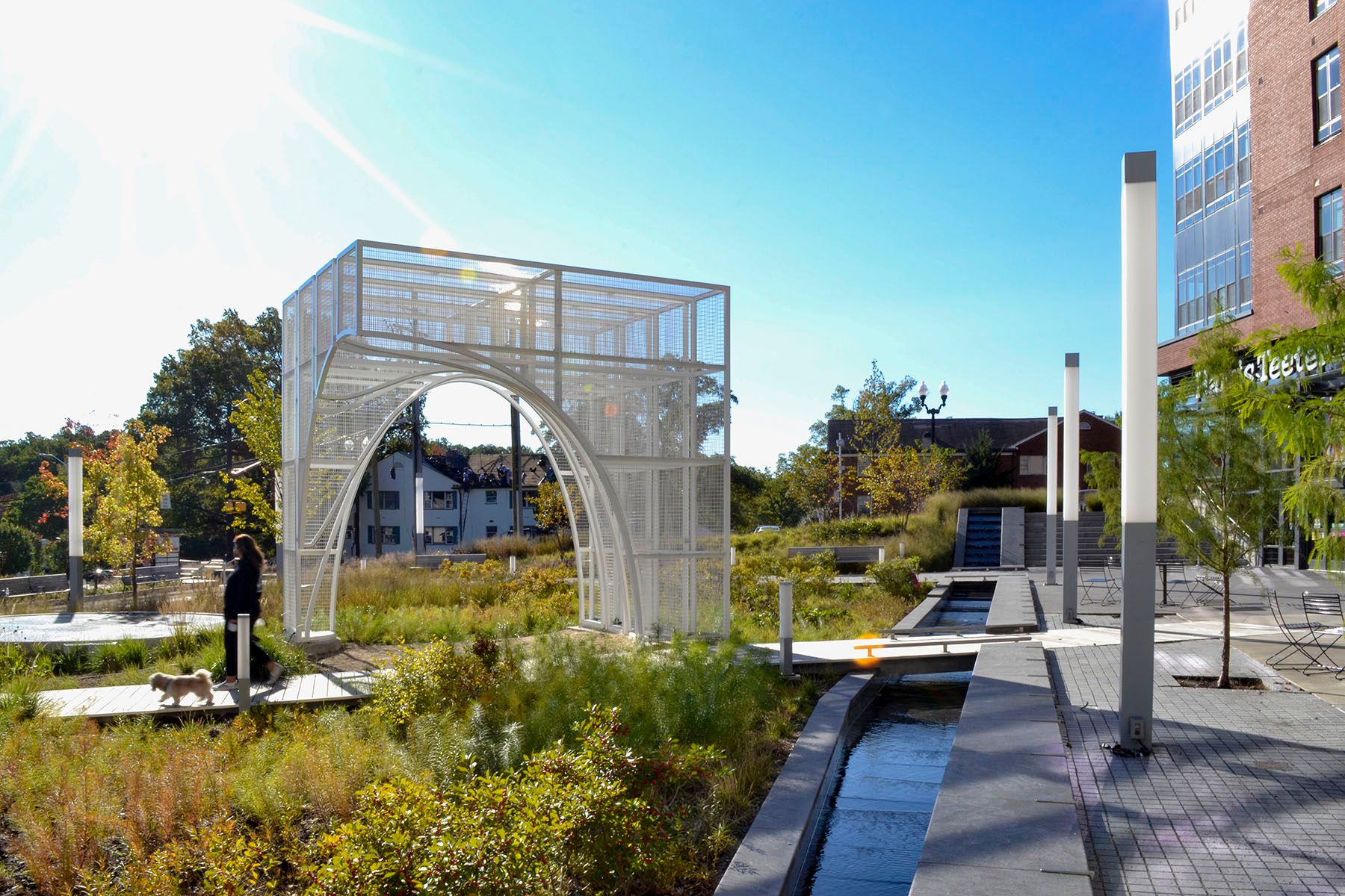 A photograph of a person with a dog walking through a very green public square, featuring a cube-like, white metal structure.
