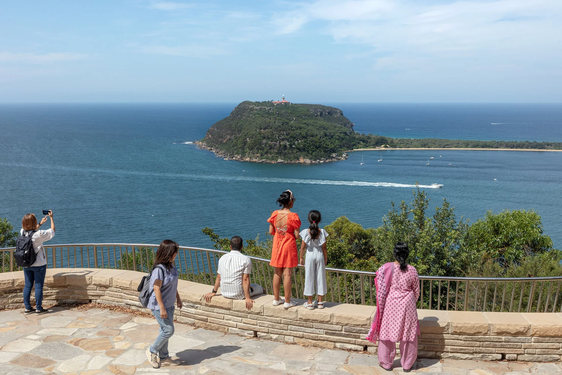Six people taking in the water views at a sandstone lookout. Two are standing on a low wall, one is sitting on the wall, one is standing behind the wall taking a photo.