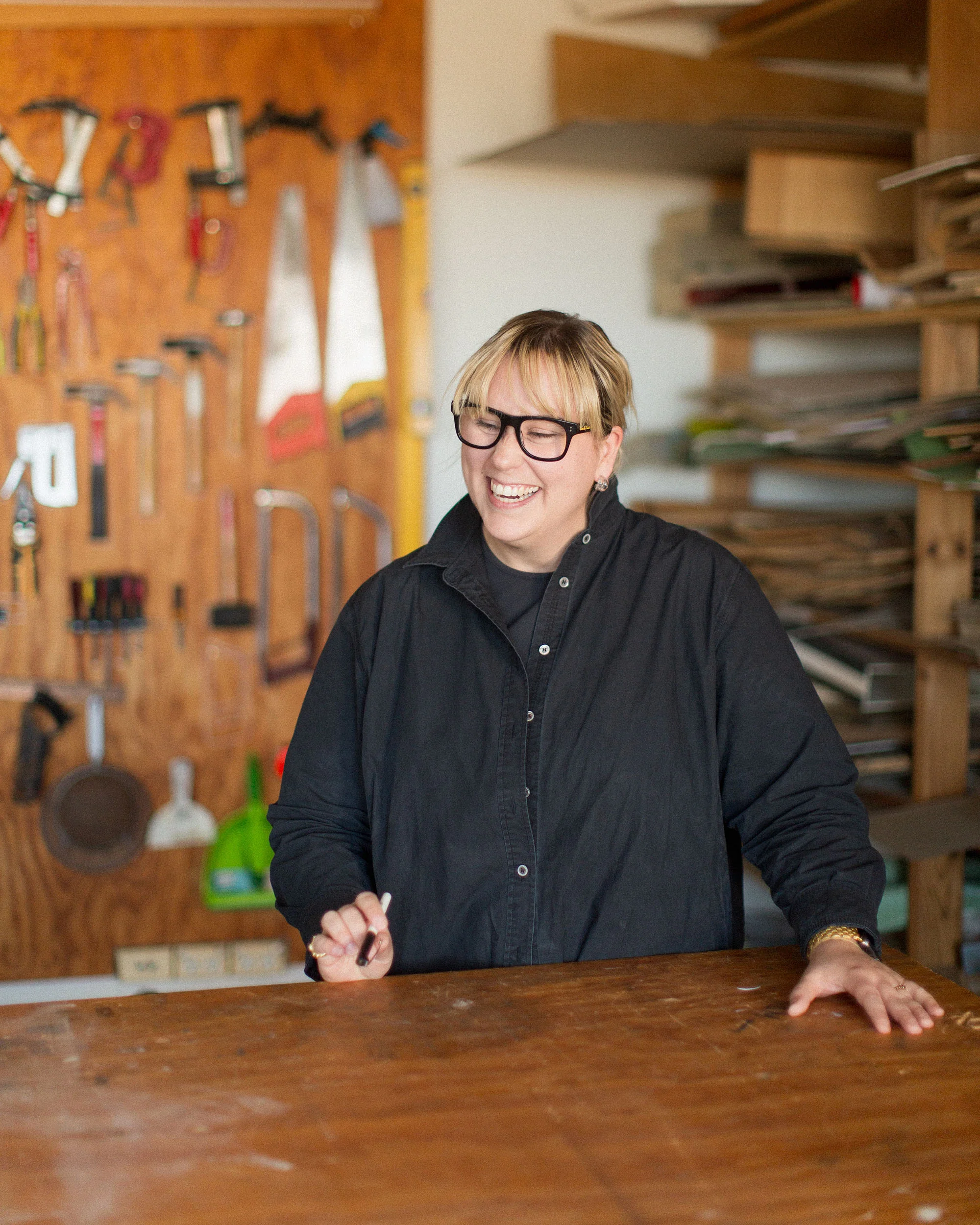 A photograph of a person laughing. They are wearing a black shirt and black glasses, standing behind a timber table. There are tools on the wall behind.