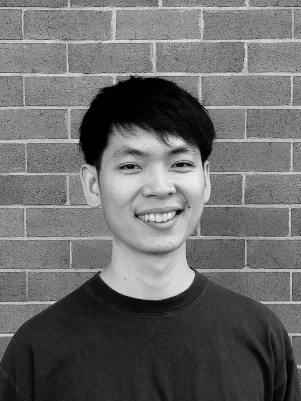 A black and white headshot of a smiling person in front of a brick wall.