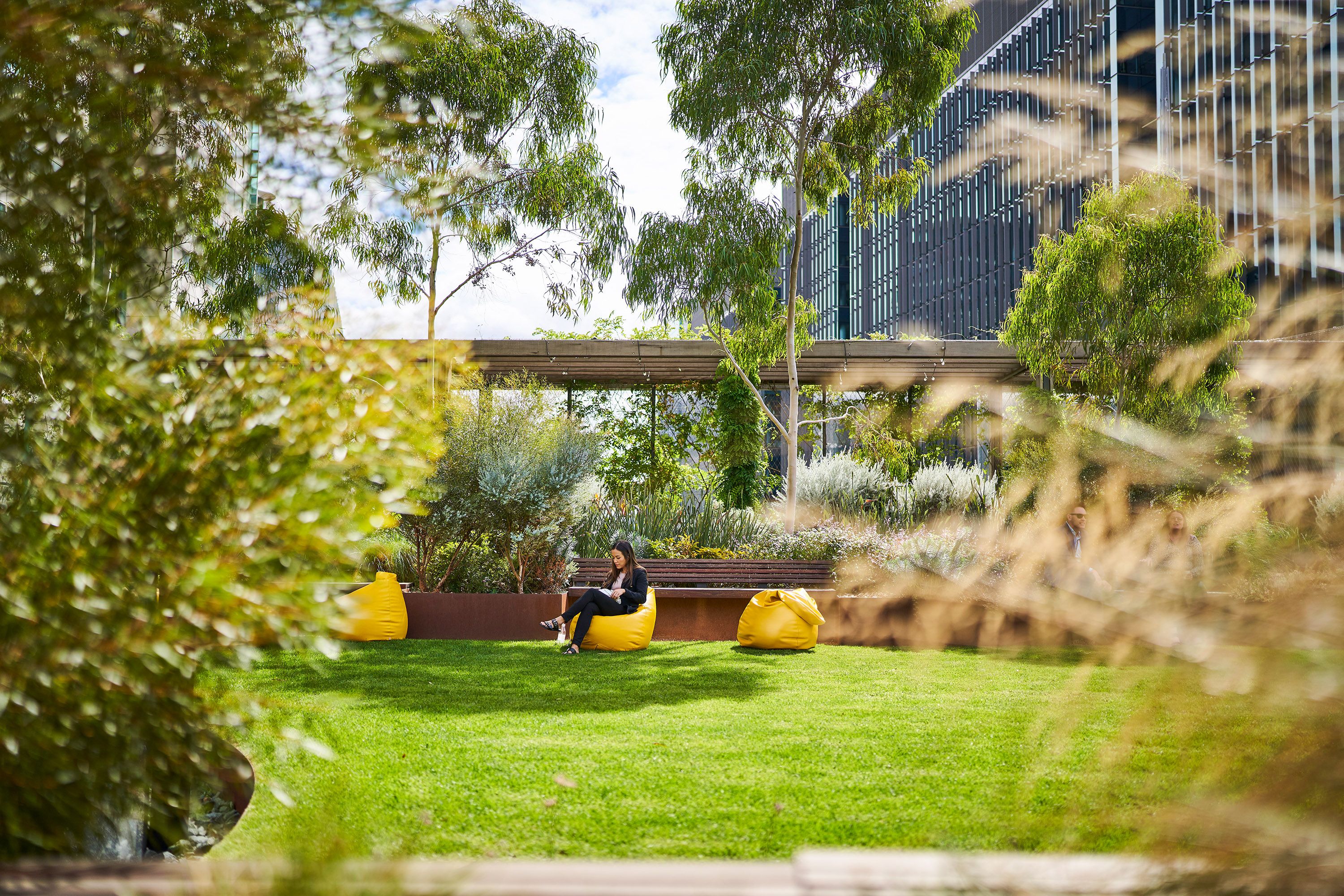 A photograph of an elevated urban park full of lush vegetation. There is a person sitting on one of three yellow bean bag chairs.