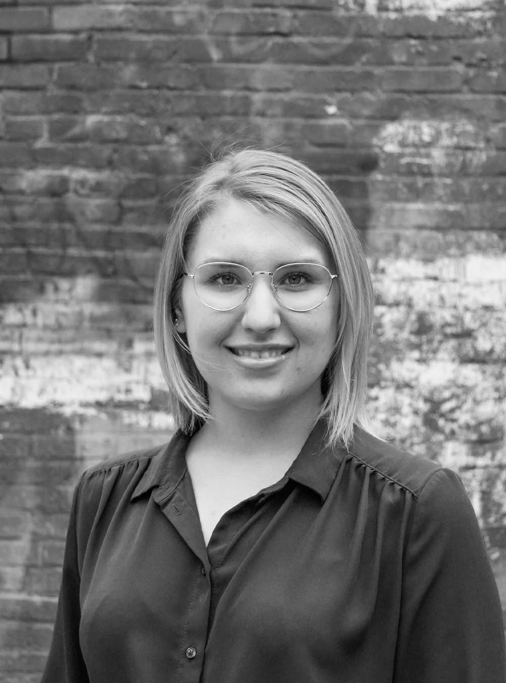 A black and white headshot of a smiling person wearing glasses in front of a brick wall.