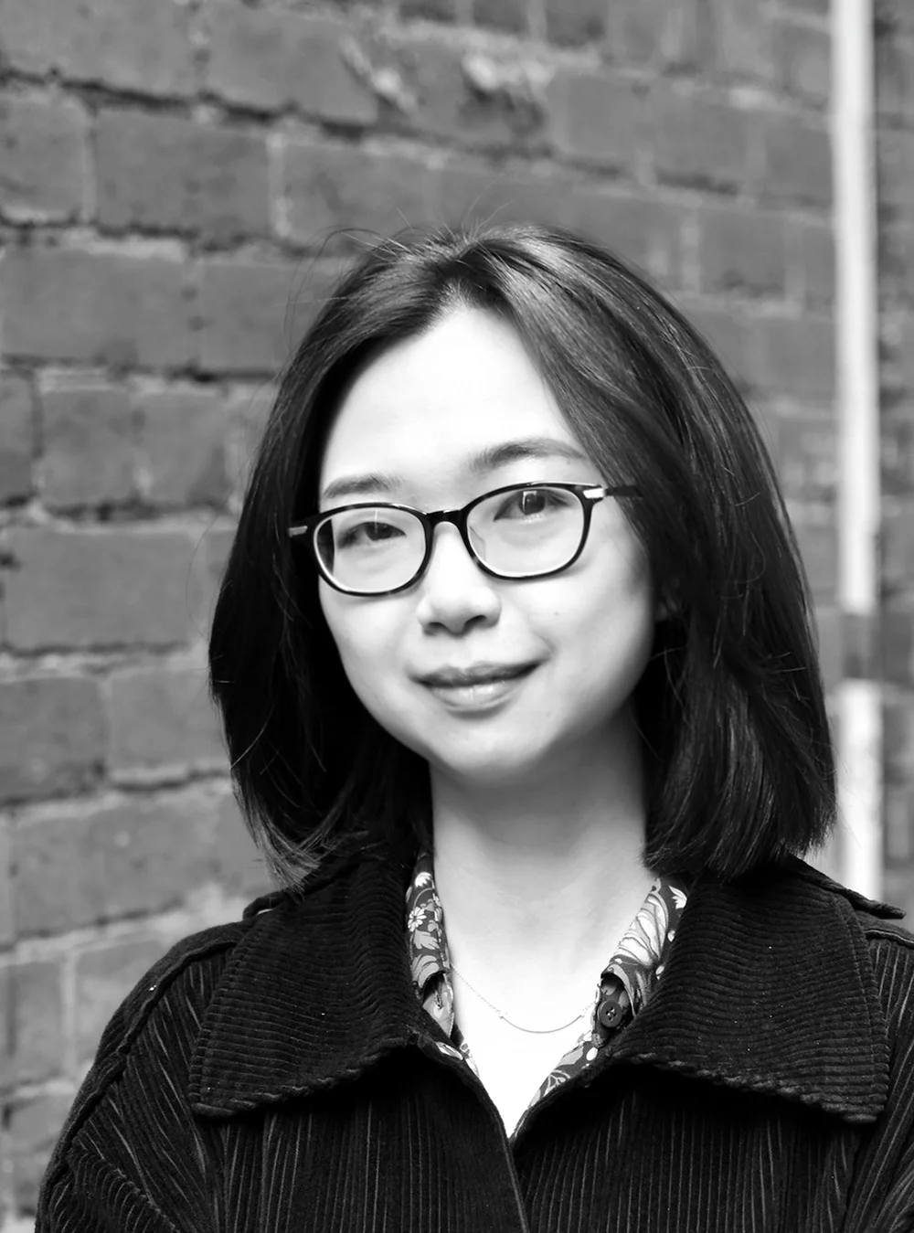A black and white headshot of a smiling person wearing glasses in front of a brick wall.