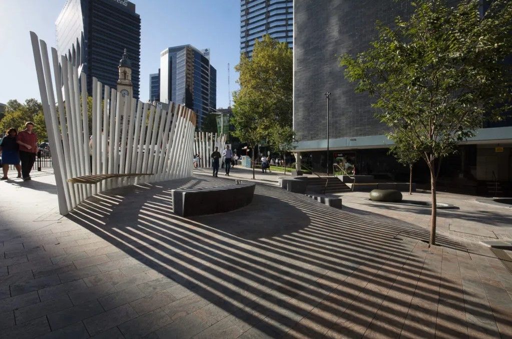 A photograph of a public plaza, there is a sculpture to the left of the image, casting long shadows across a seating area. Commercial buildings can be seen in the background