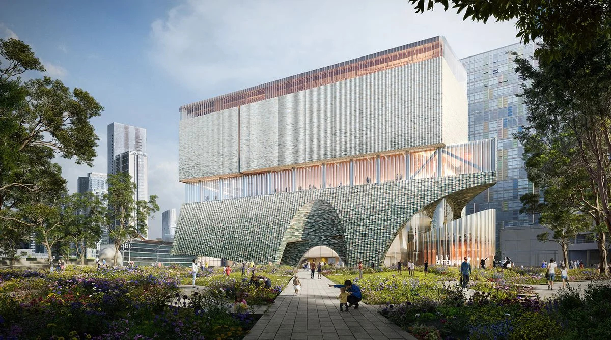 A photorealistic visualisation of a contemporary art gallery seen from ground level. It is a square form, with curved entrances and awnings at the base of the building. Trees and plants are visible in the foreground. City buildings are visible in the background.