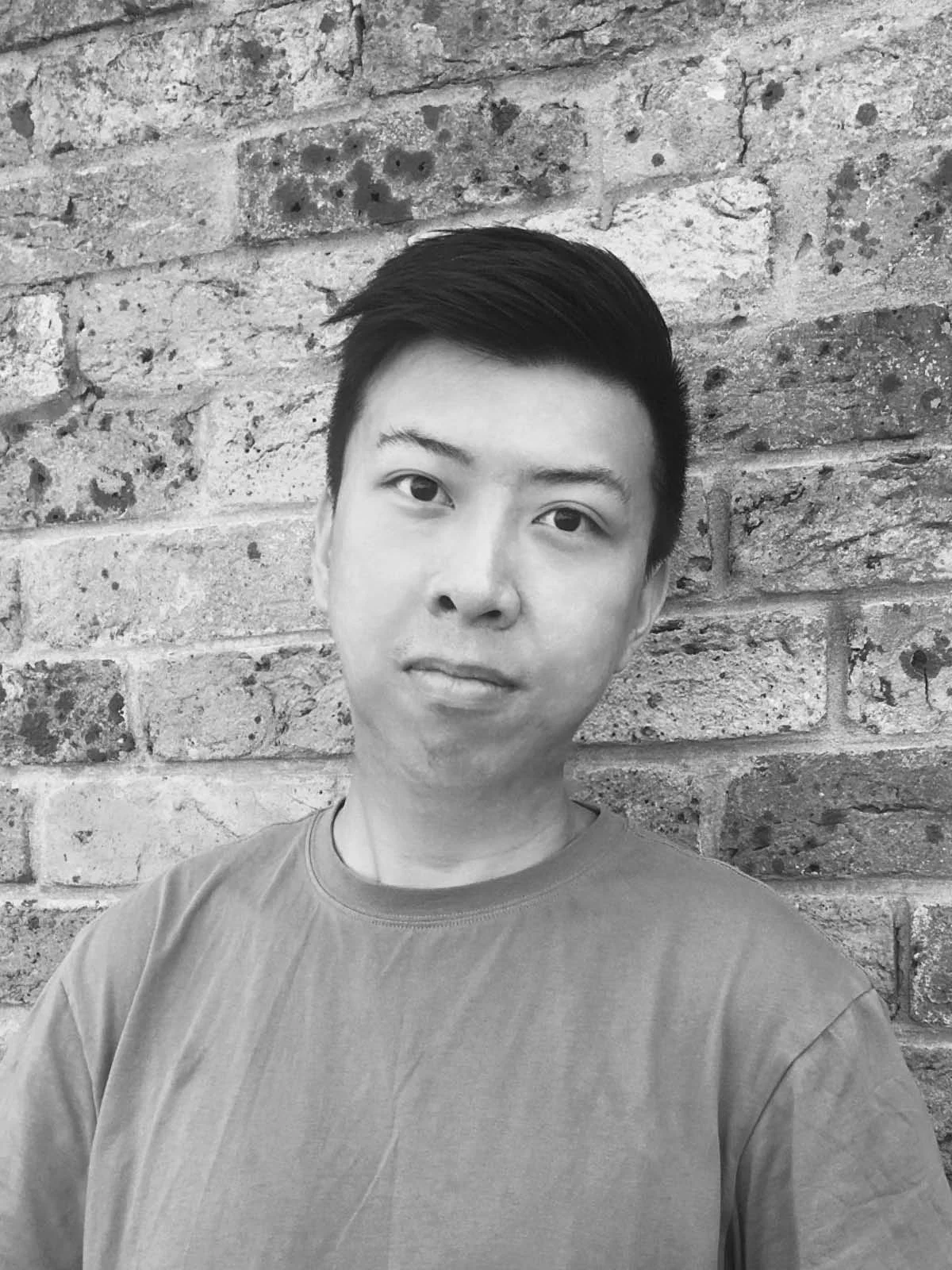 A black and white headshot of a person wearing a t-shirt, in front of a brick wall.