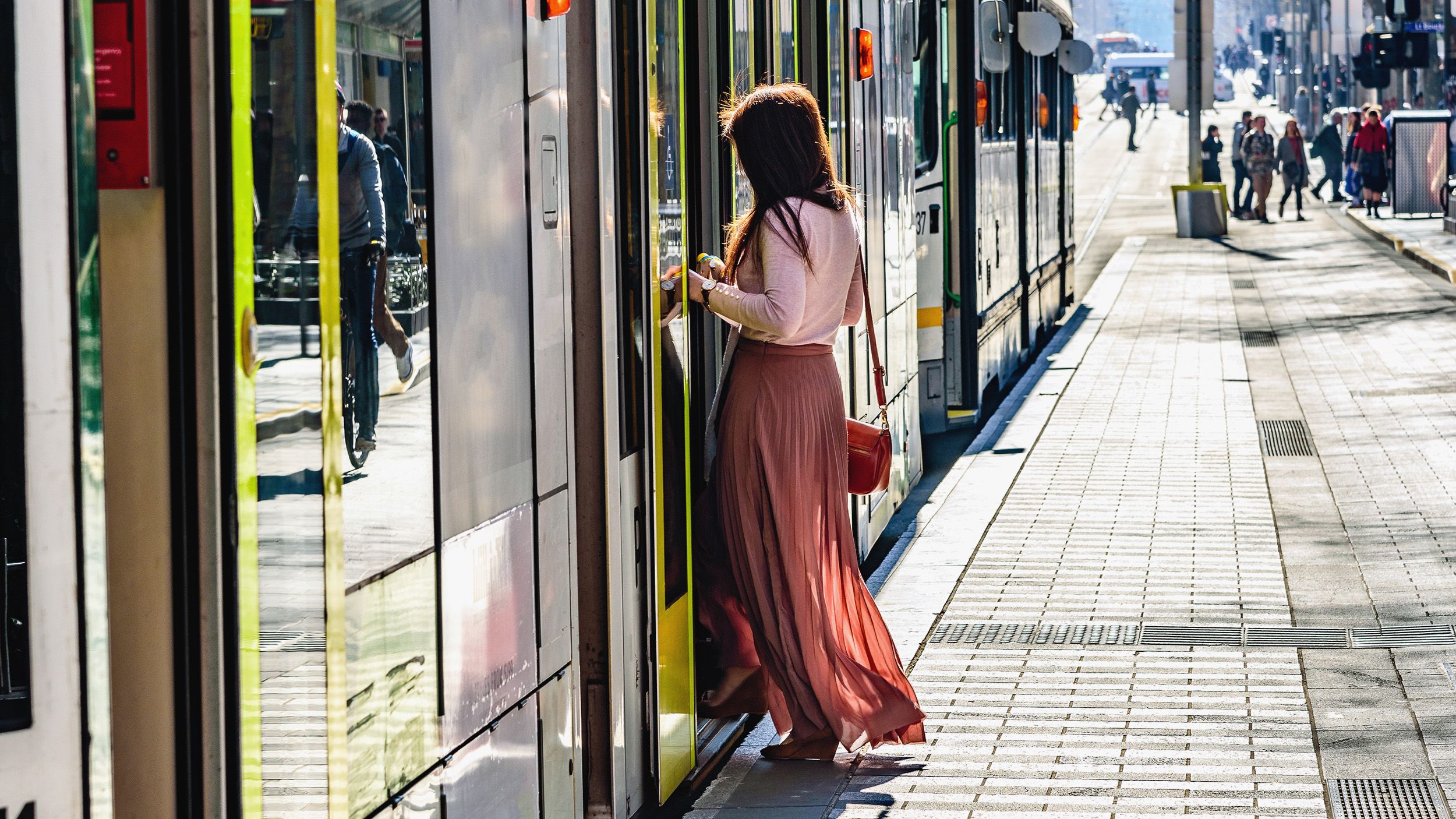 A photograph of person in a long flowing skirt stepping onto a tram in urban Melbourne. There are other people in the distance, and a reflection of a cyclist in the tram doors.