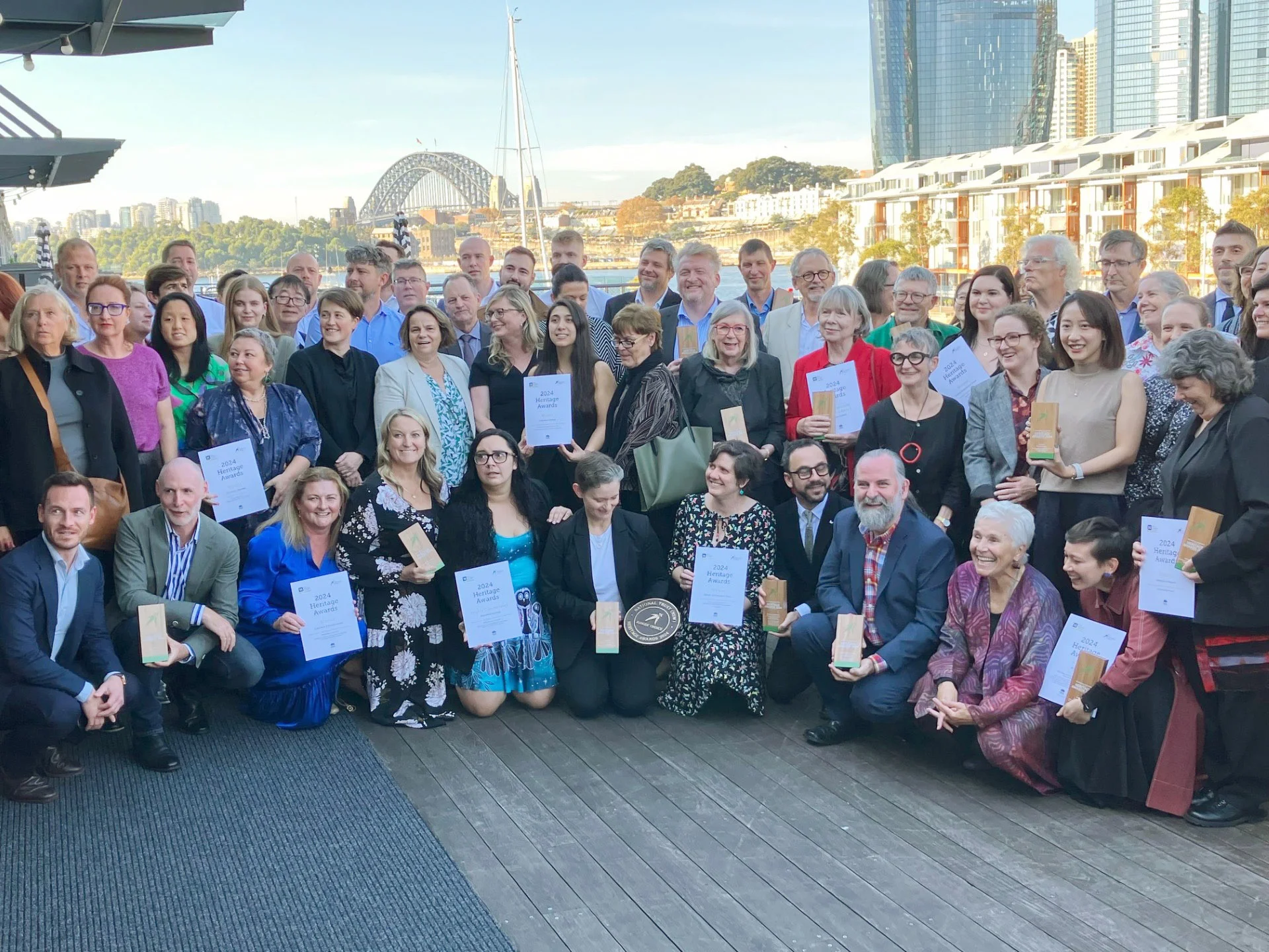 A photo of a large group posing with certificates and trophies. The Sydney Harbour Bridge is in the distance behind them.