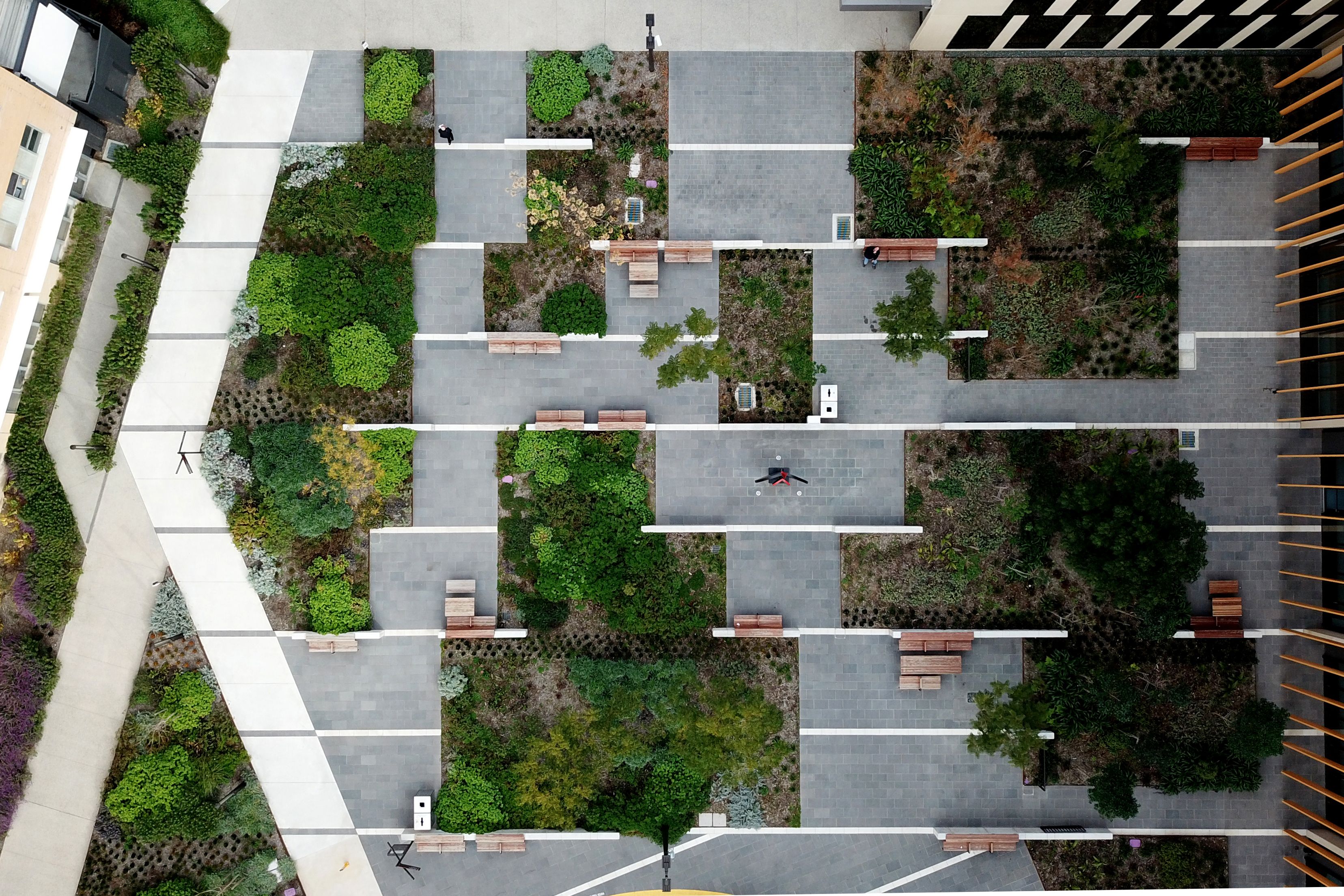 A drone photograph of large public courtyard. It's a grid of grey paved and green planted areas, with a white angled path on the left.
