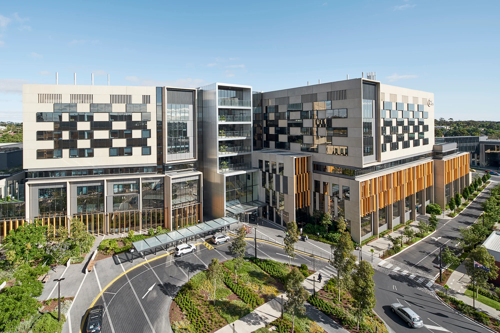 A photograph of a new hospital building. There is a road and drop off circle in the foreground, as well as tree lined pathways and a lush, green forecourt.