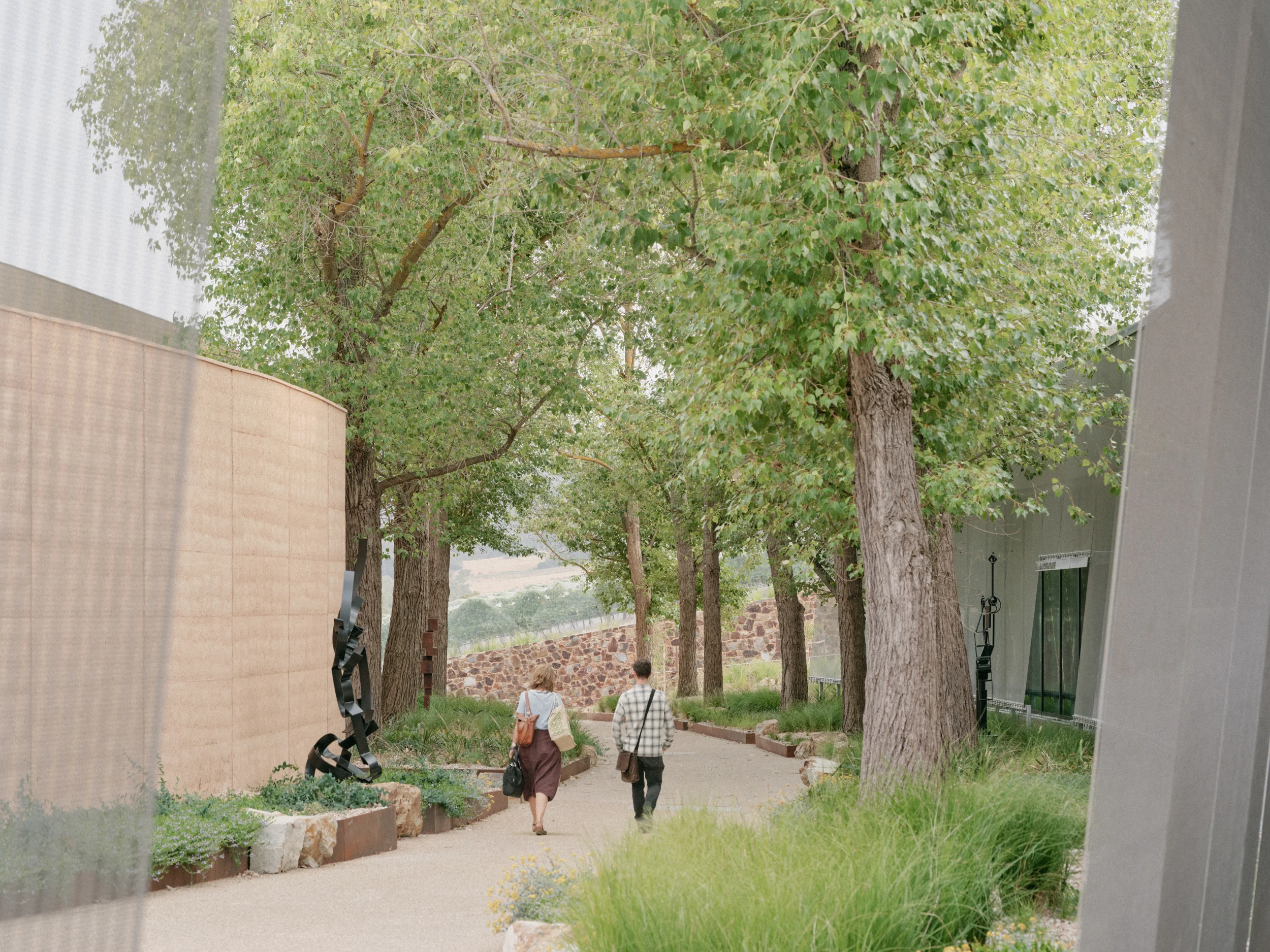 A photo of two people walking through a curved outdoor passage between buildings, past a sculpture towards an avenue of trees. They are both carrying bags.
