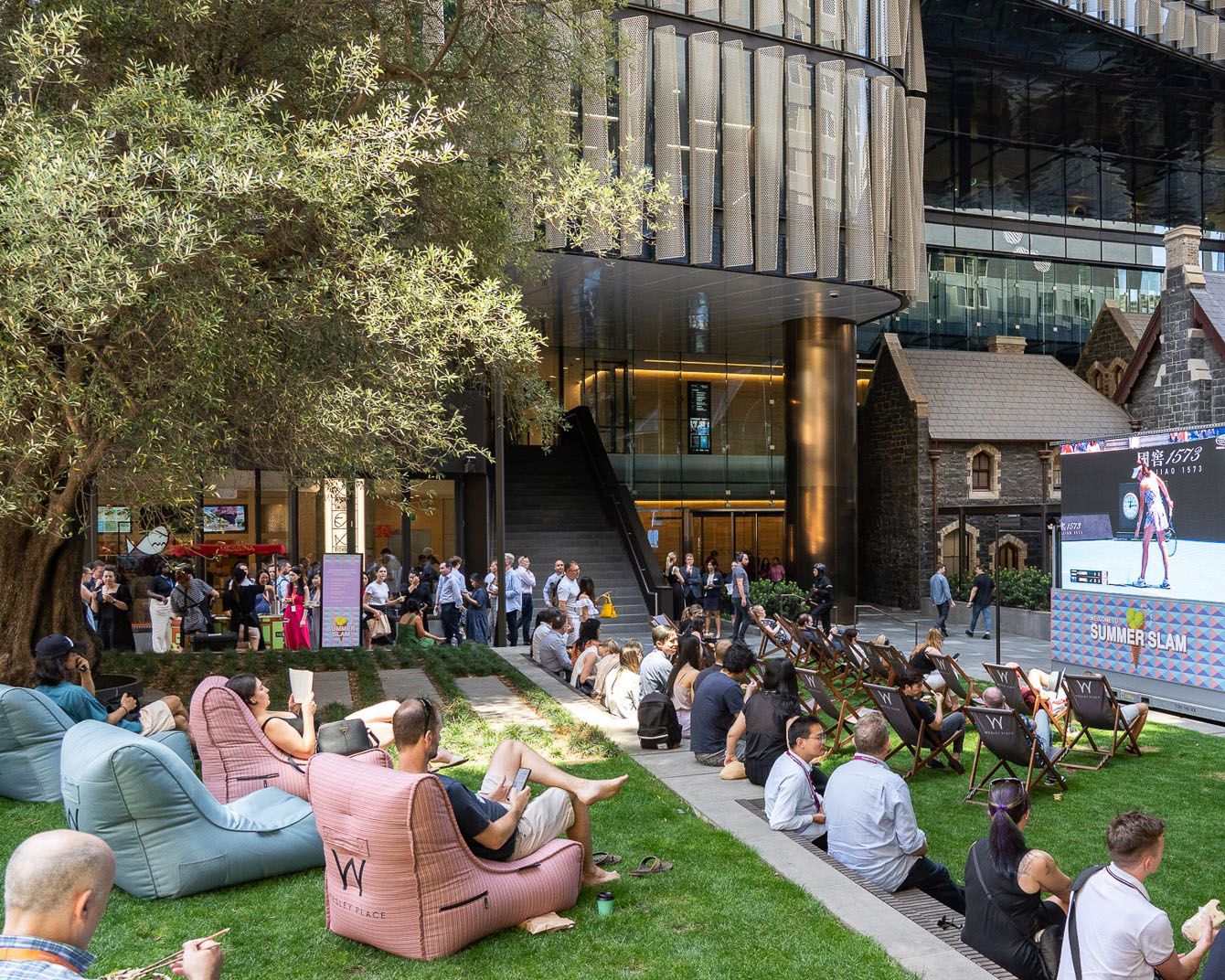 A photograph of a public, urban, green courtyard filled with people sitting and watching a tennis game on a large screen.