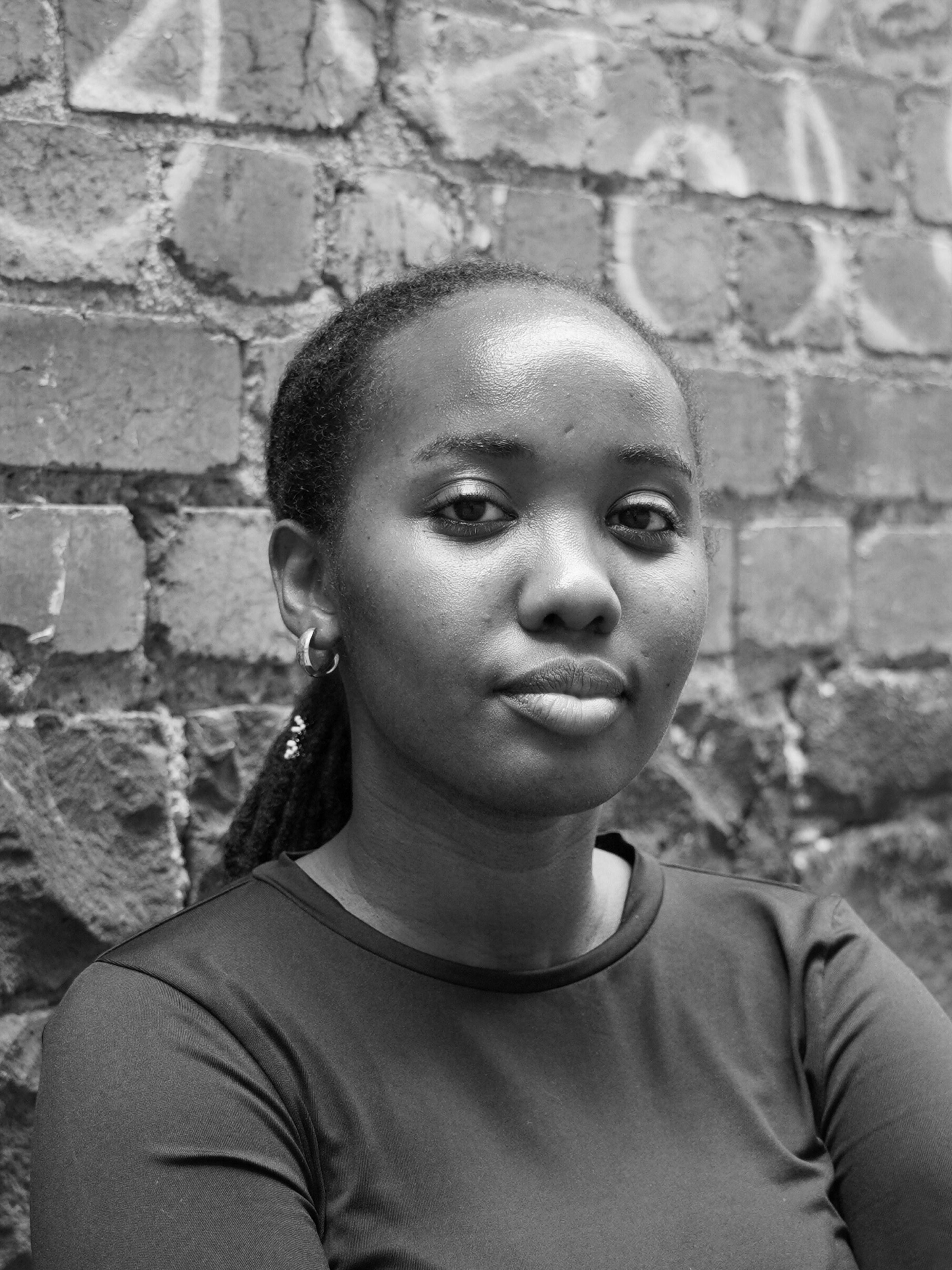 A black and white headshot taken in front of a graffitied brick wall.