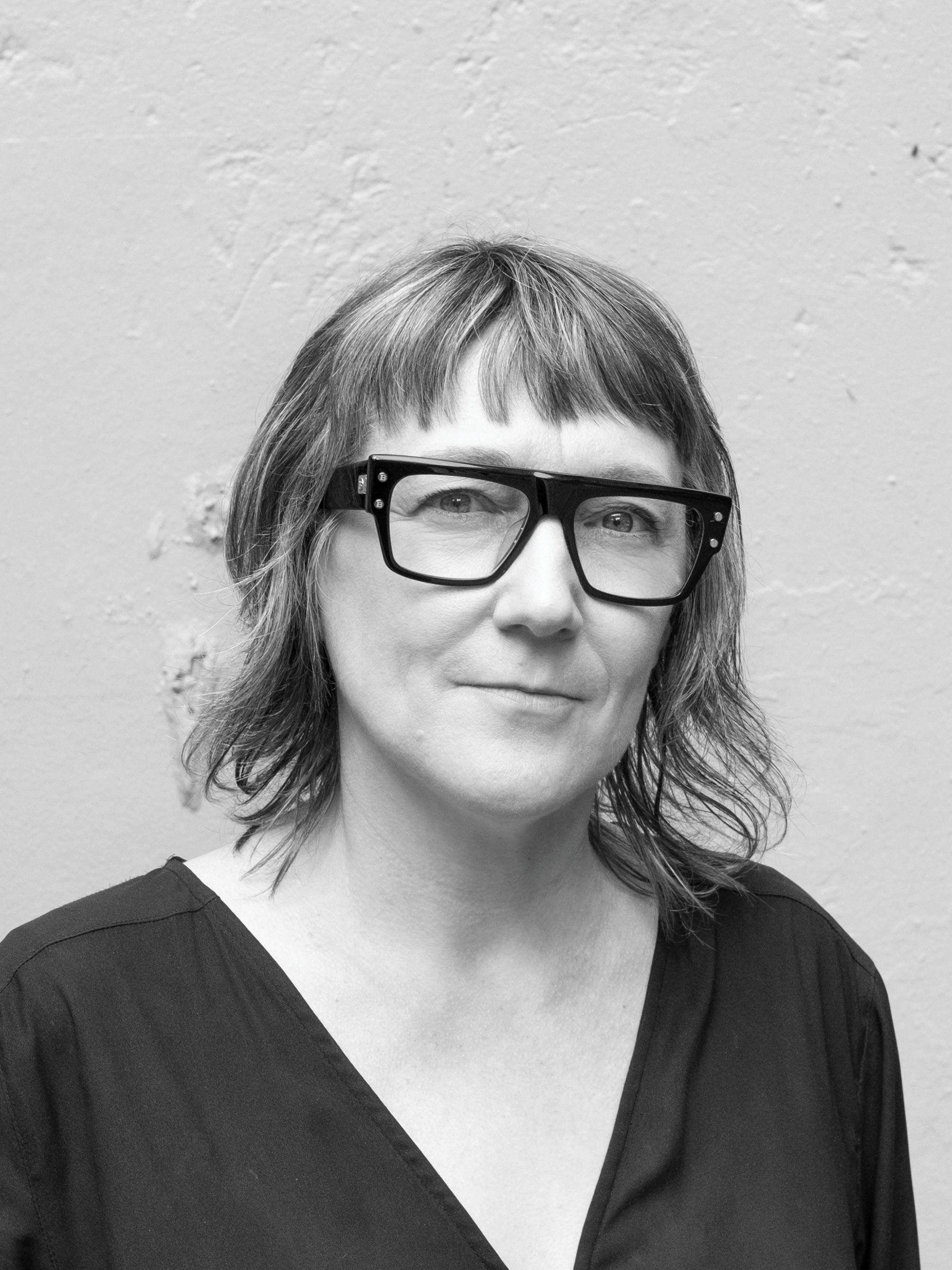 A black and white headshot of a person wearing distinct black glasses and standing in front of a painted outdoor wall. 