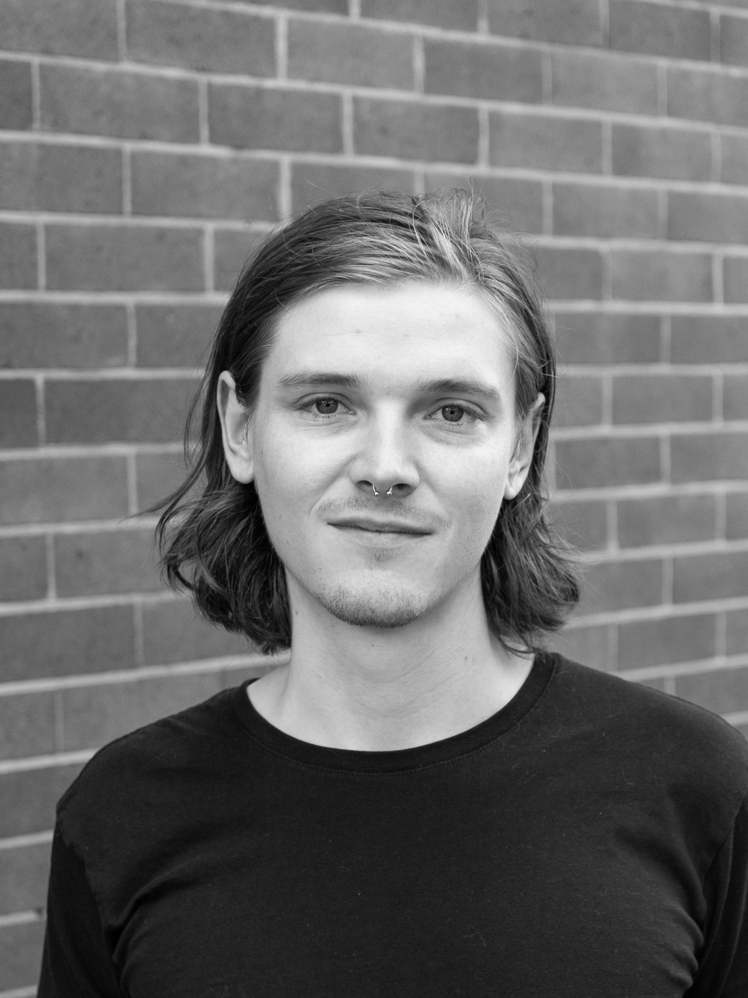 A black and white headshot of a person in front of a brick wall.