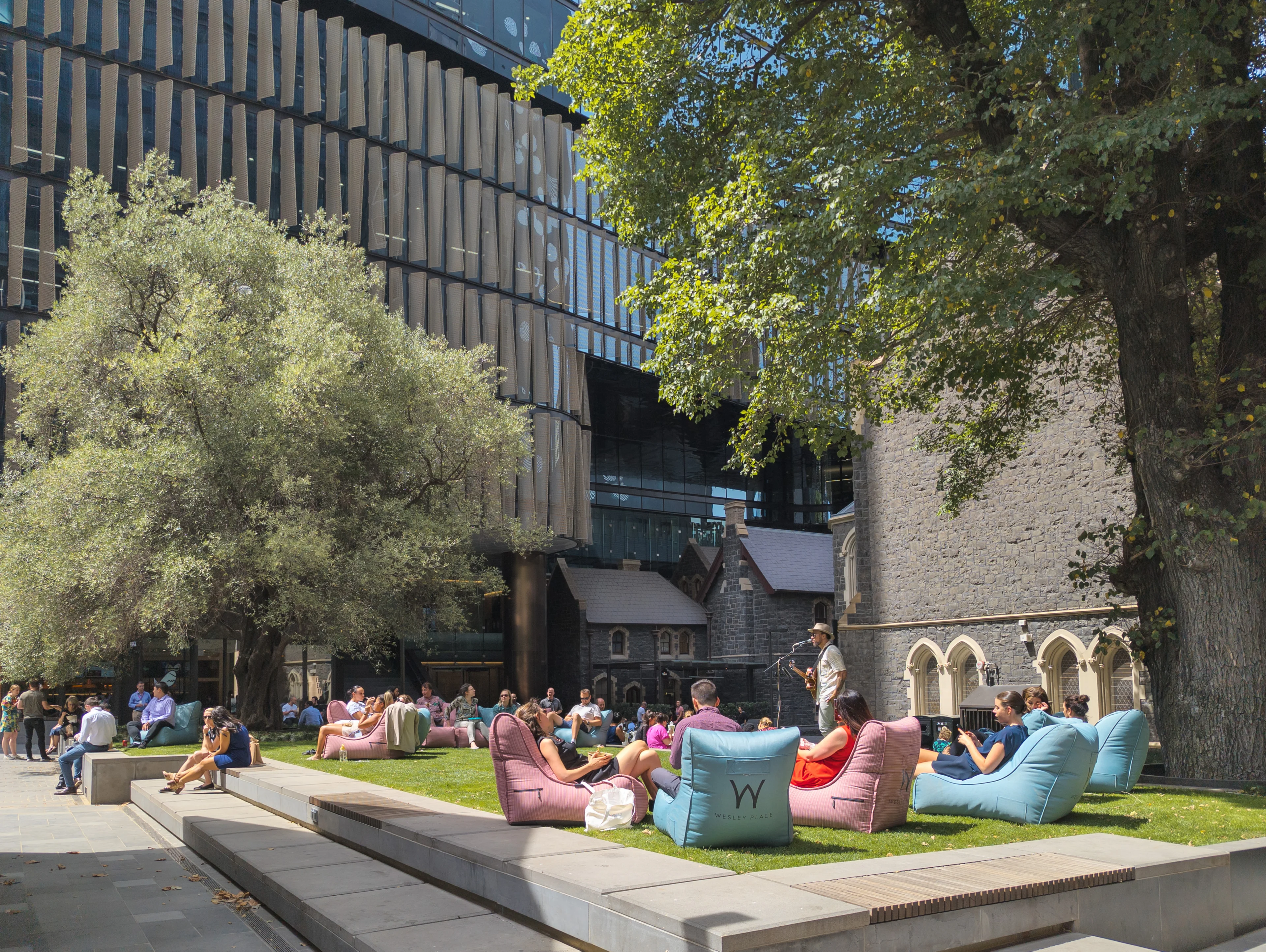 A photograph of an urban green square, there are two large trees, and a crowd of people sitting on pink and blue bean bag chairs listening to a live musician.