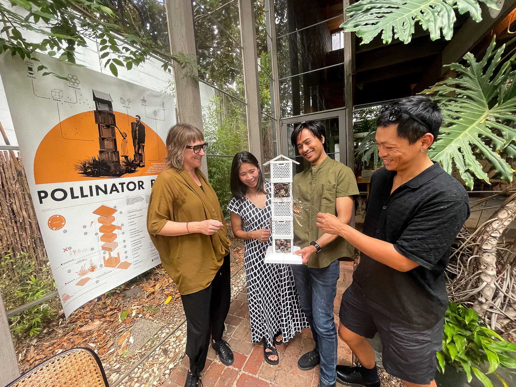 A photo of four smiling gathered around a geometric white model filled with dry organic material like twigs and seed pods. The photo was taken in a courtyard.