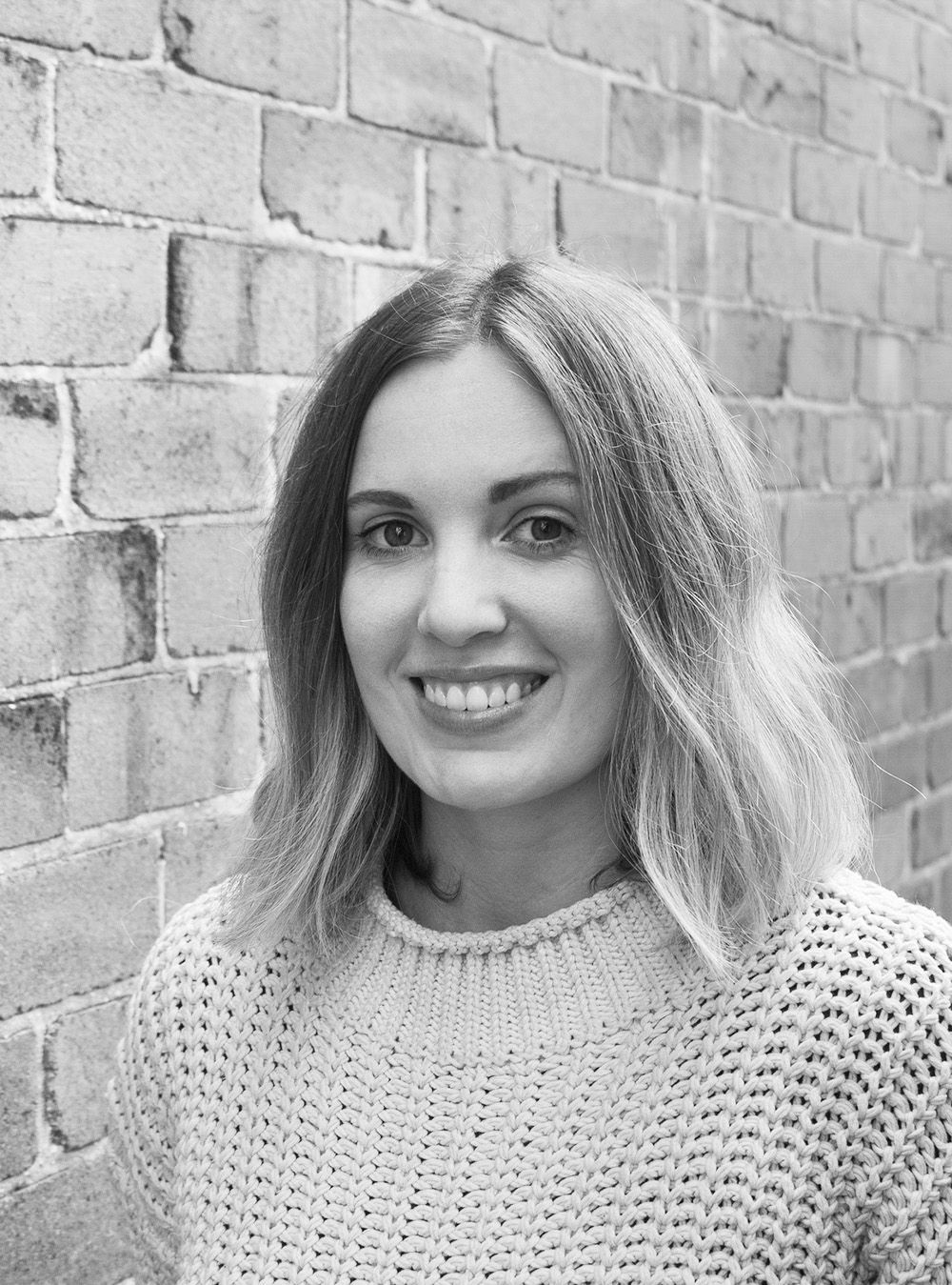 A black and white headshot of a smiling person in front of a brick wall.