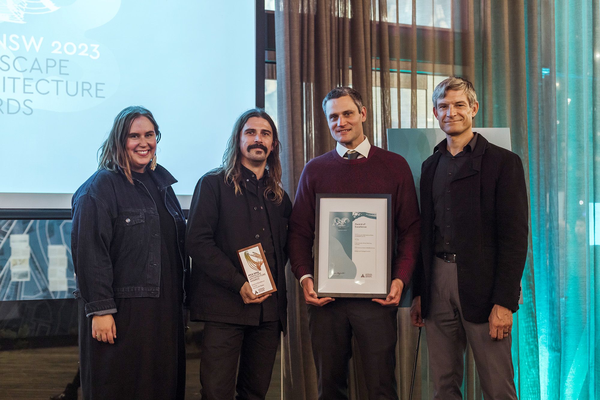 A photograph of four people on an awards stage, holding a small square trophy and a framed certificate. 