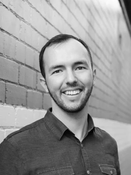 A black and white headshot of a person smiling in front of a brick wall.