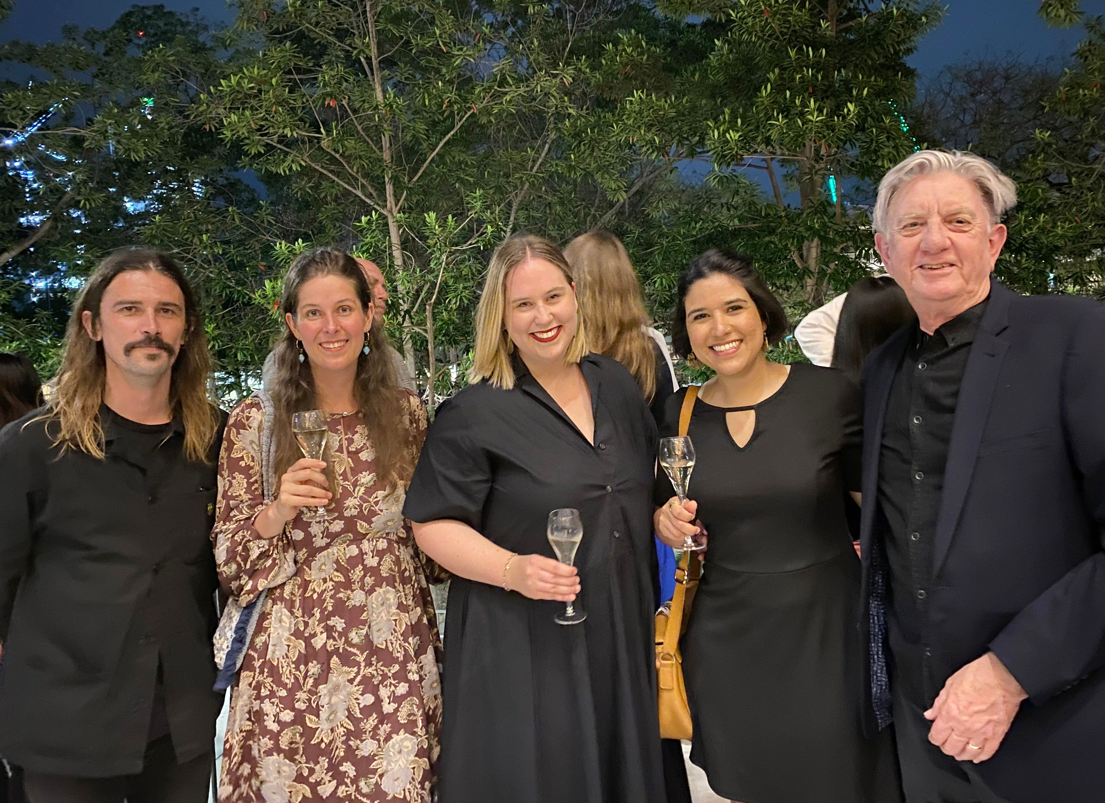 A photograph of five people in a row, smiling at the camera.  Four are wearing dark colours, three are holding wine glasses, two have short hair. Above their heads we can see trees and a night blue sky.