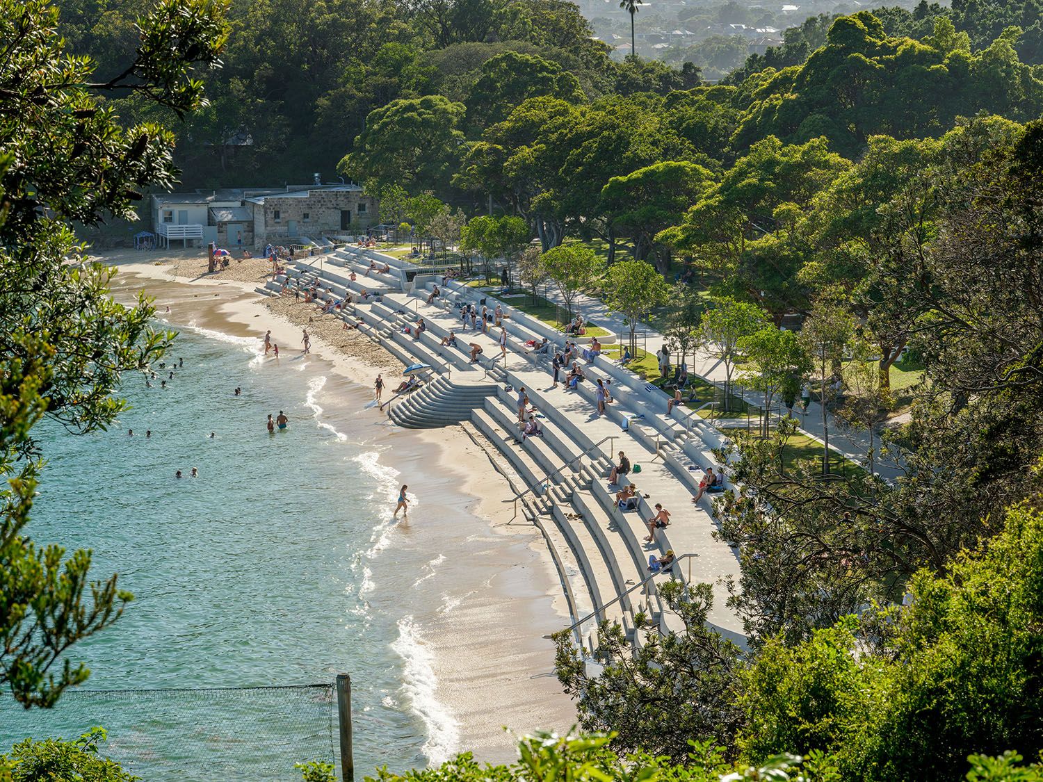 A photograph taken looking down the length of a beach seawall on a sunny day. The image is framed by green trees. 